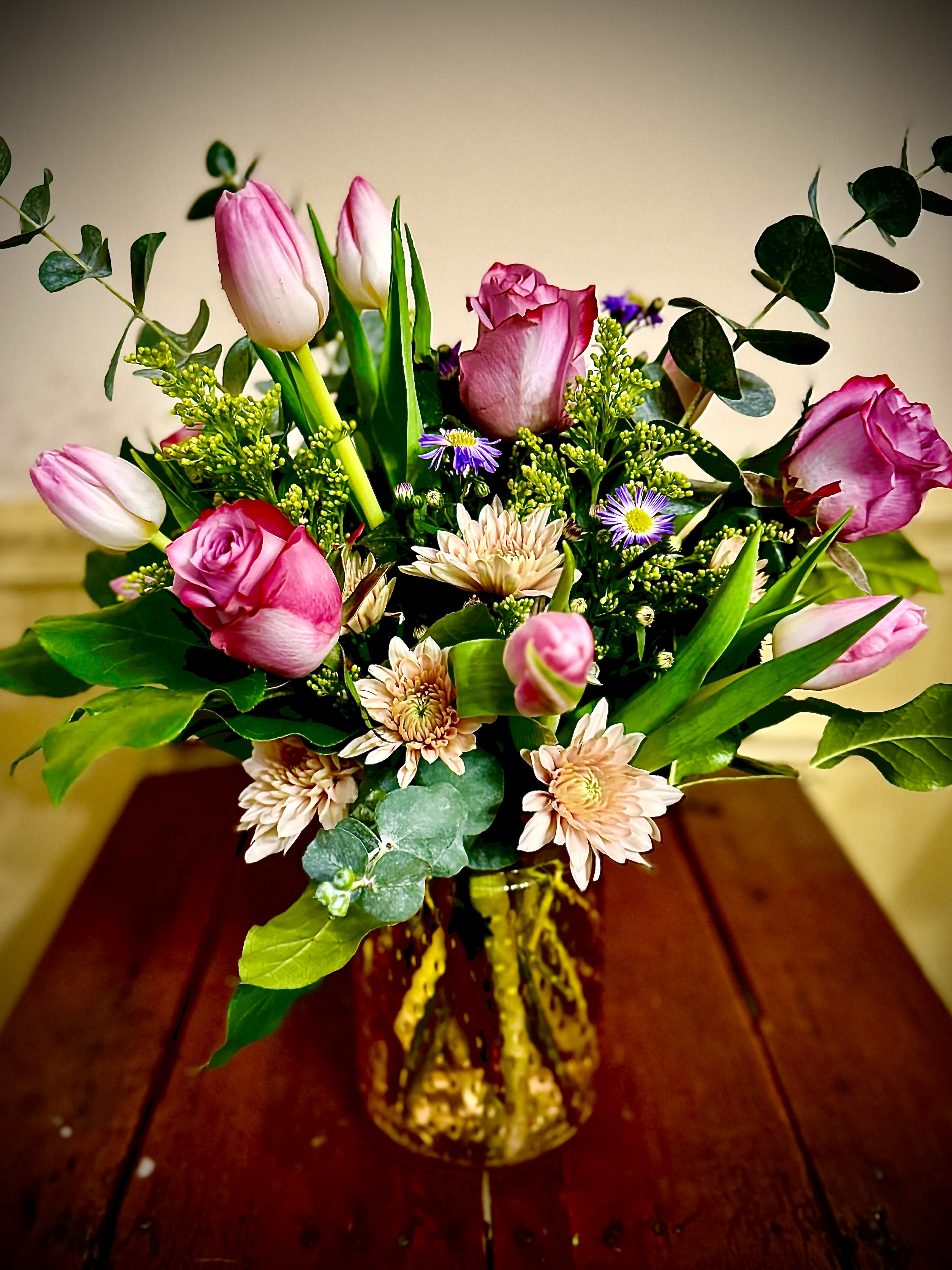 Pink roses, tulips, and pale peach mums with purple accents in an amber glass vase.