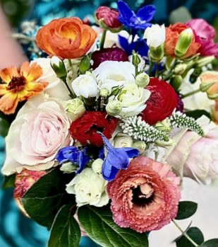 Colorful hand-tied bouquet with orange ranunculus, blush roses, red blooms, and blue accent flowers.