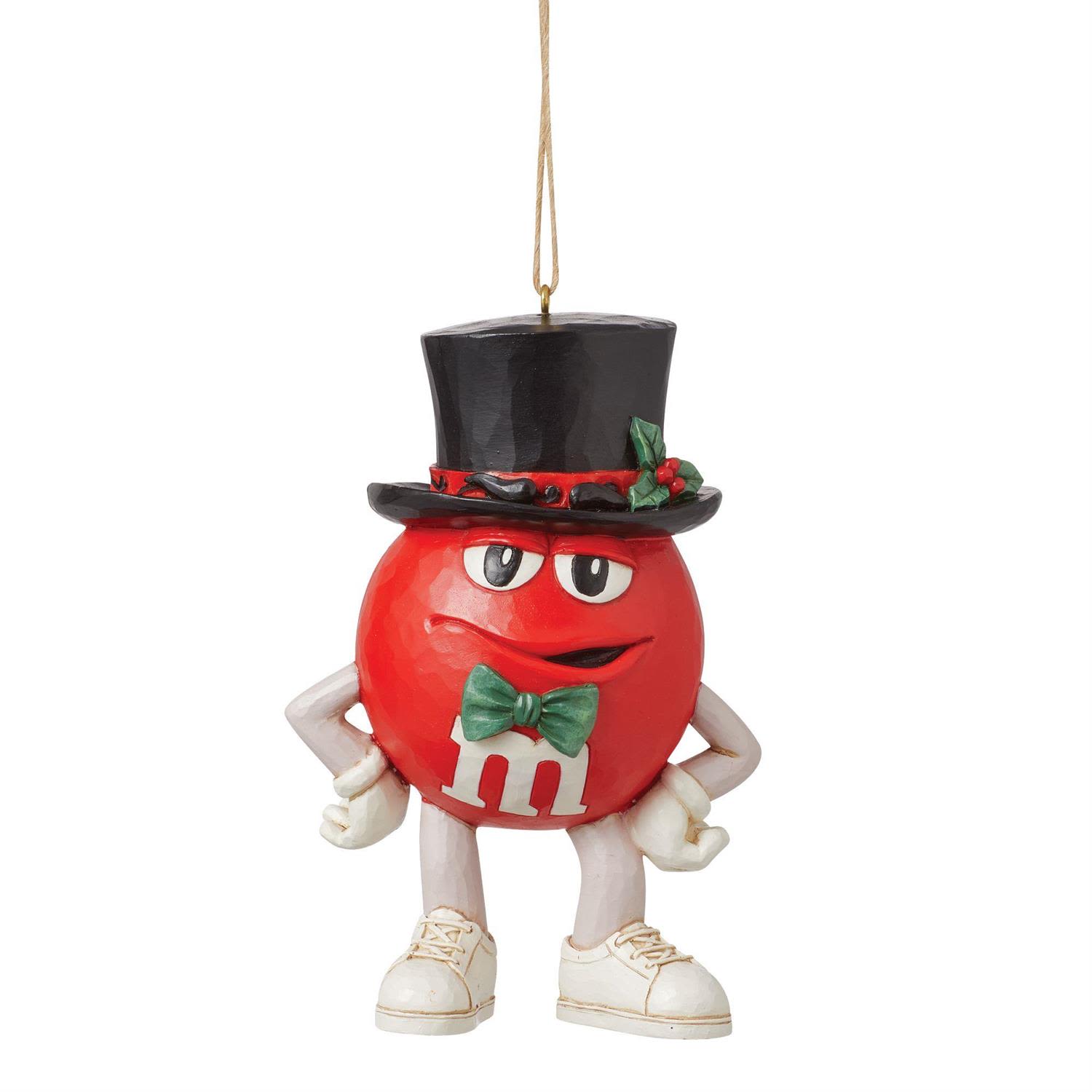 M&amp;M’S Red Character in Top Hat Ornament - Make room for a little charm—and a lot of attitude—with the M&amp;M’S Red Character in Top Hat Ornament. Known for his confident (and occasionally self-absorbed) personality, Red is dressed to impress this season. Wearing an elegant top hat, a festive bow tie, and a sprig of mistletoe, he’s fully prepared to charm his way through the holidays with his signature mix of chocolate and cheer.  Standing 4.33 inches tall, this hanging ornament is beautifully hand-painted and crafted from high-quality stone resin, showcasing the intricate details and nostalgic motifs that define the artistry of Jim Shore. From subtle folk-art patterns to classic quilt-inspired textures, Red’s festive look blends playful character design with timeless holiday craftsmanship.  Each ornament is sold individually and available in limited quantities, making it a delightful addition for collectors, M&amp;M’S fans, and anyone who loves a touch of personality on their Christmas tree.
