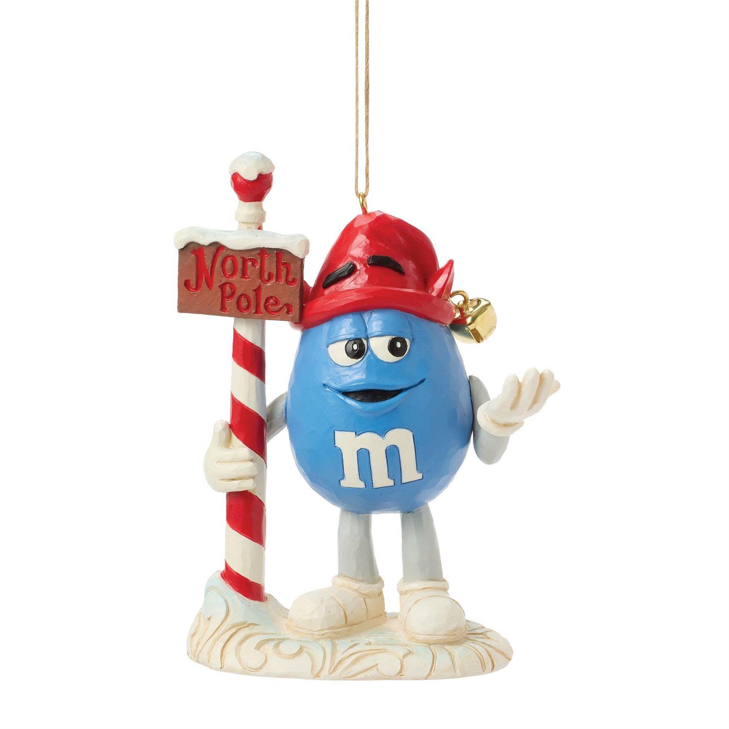 M&amp;M'S Blue Character Elf Ornament - Bring a dash of cheeky charm to your holiday décor with the M&amp;M’S Blue Character Elf Ornament from the Jim Shore Collection. Ever the cool and carefree personality, Blue stands proudly beside a festive North Pole sign, dressed in a bright red elf hat and wearing his signature playful smirk. With a jingle bell at his cap and an easygoing wave, he’s ready to spread holiday mischief and chocolate cheer all season long.  Standing 4.65 inches tall, this hanging ornament is beautifully hand-painted and crafted with the intricate folk-art patterns and nostalgic detail that define Jim Shore’s style. The carved textures, whimsical base, and rich color palette make this piece both collectible and irresistibly fun.  Each ornament is sold individually and available in limited quantities, making it a delightful addition for M&amp;M’S fans, Jim Shore collectors, or anyone who loves a touch of personality on their Christmas tree.