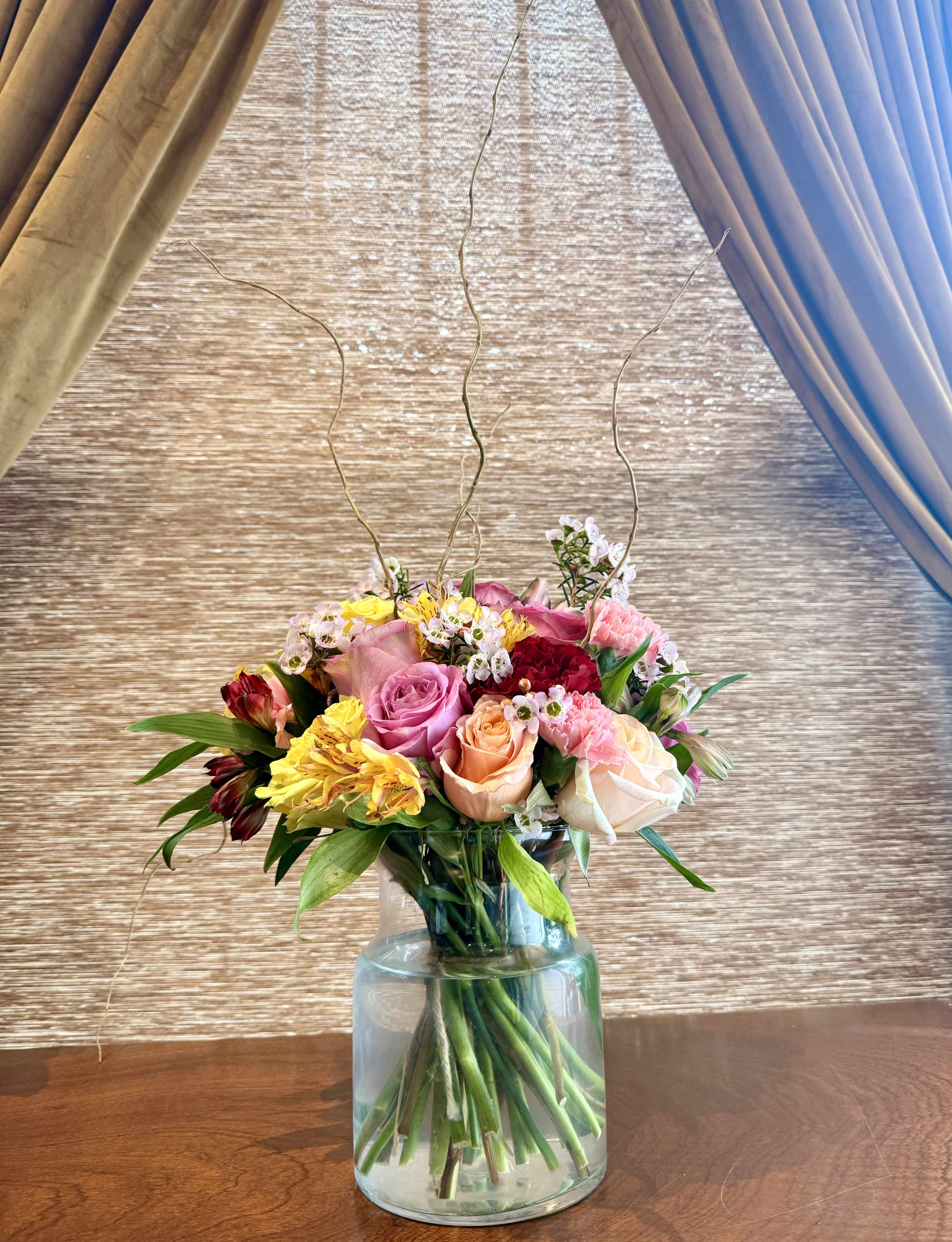 The Story We Tell Arrangement - Soft, poetic, and deeply felt.  Designed as a modern garden composition, this arrangement highlights premium garden roses—including Pink Expression and Phoenix—chosen for their nuanced tones and luxurious petal structure. Sculptural protea introduces gentle structure and depth, while soft accents and waxflower create a delicate, meadow-like texture. Branching elements rise above the blooms to add narrative, lift, and elegance. Natural, whimsical, and refined, it feels like a love story unfolding—quietly dramatic, never overstated.  PART OF OUR LOVE IN EVERY ERA — ARRANGEMENT COLLECTION Featuring Say It Out Loud, The Story We Tell, and Midnight Moment  This Valentine’s collection is intentionally bloom-forward, designed around a tightly curated selection of premium garden rose varieties chosen for their exceptional petal quality, fragrance, and presence. To honor the integrity of each design, we are limiting our Valentine’s stock to these featured blooms, allowing the roses — and their natural beauty — to take center stage. Our arrangements are lush, expressive, and refined, accented with delicate waxflower, sculptural protea, and elegant branching to create movement, depth, and lasting visual impact.  Images shown are renderings and are representative of overall size, style, and color palette.
