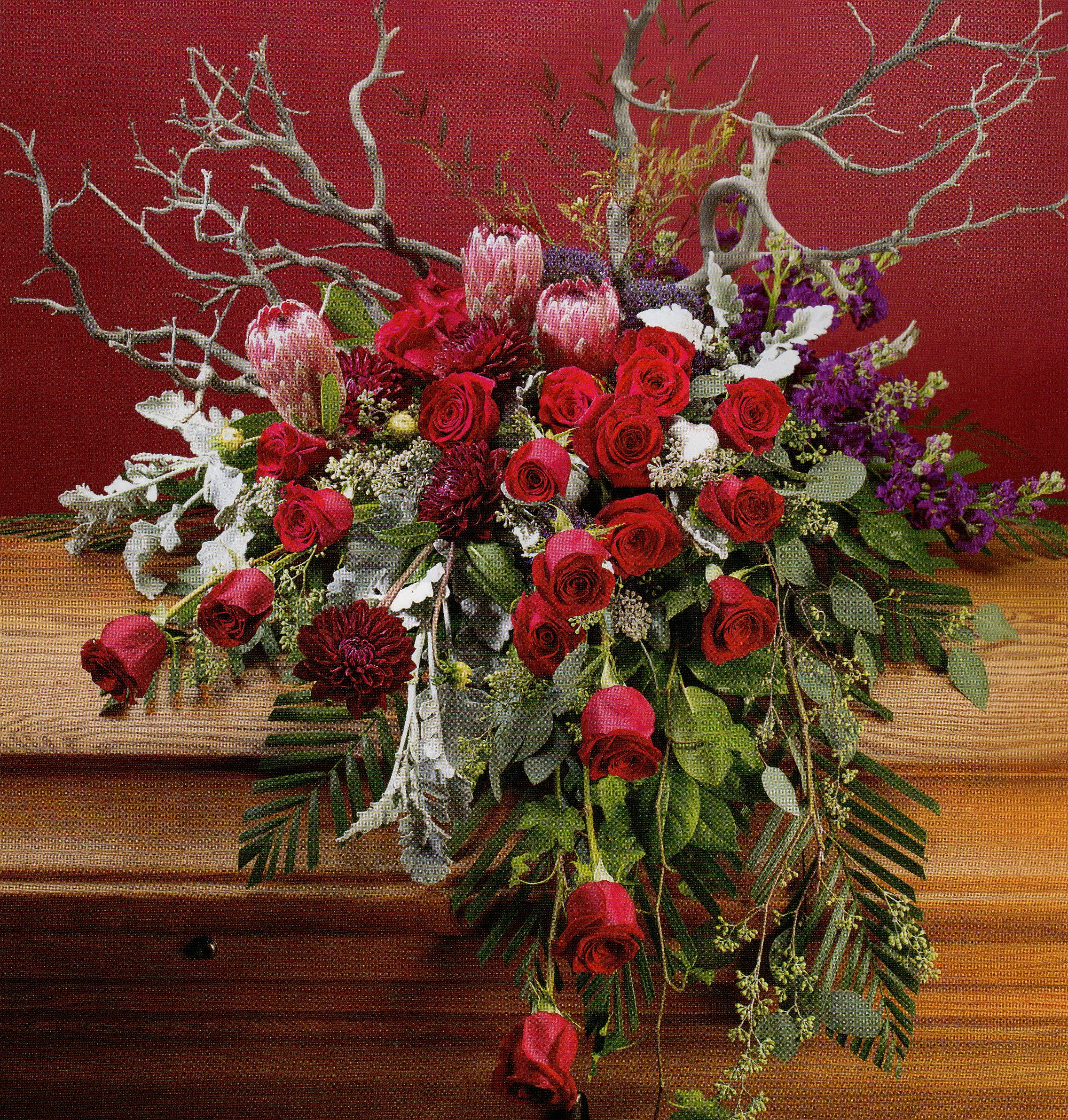 Rustic Roses Tribute Spray - This exquisite casket spray comes complete in ruby and amethyst jewel tones. Roses, spicy stock, and queen protea are accented with fine foliage texture and branches,