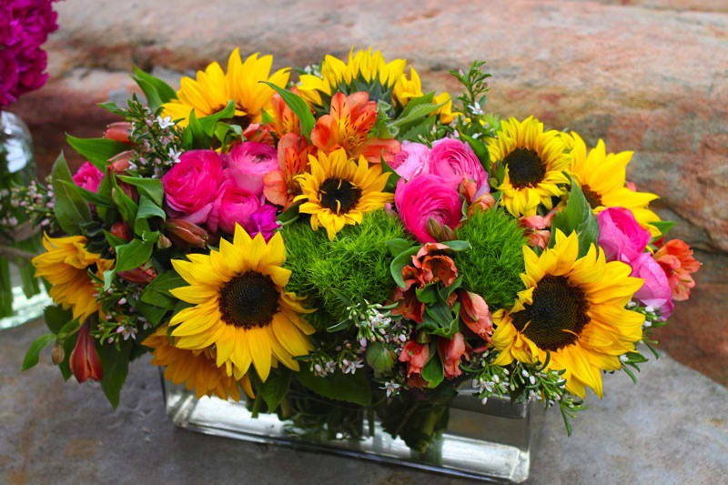 Summer Lovin' - Gorgeous Vibrant bouquet nestled in oversized rectangular clear vase and filled with Eye popping Sunflowers, Fluffly Hot pink ranunculas, Green trych, Orange alstromeria lilies, and filler....