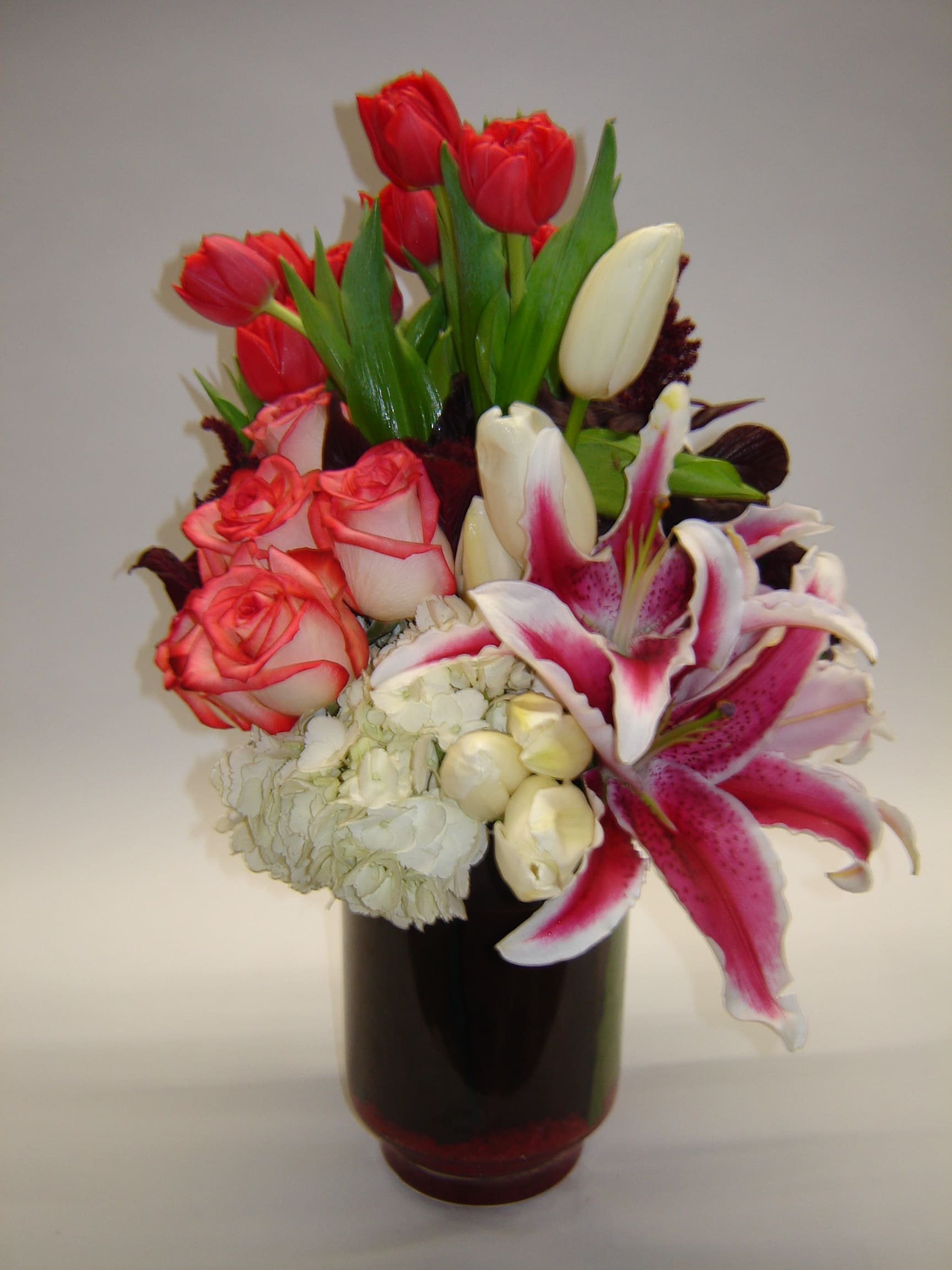 In Sync Style In Sync Style - A pave' style arrangement with tulips, roses, stargazer lilies, hydrangea complement each other in this cylinder vase lined with a plum Ti leaf