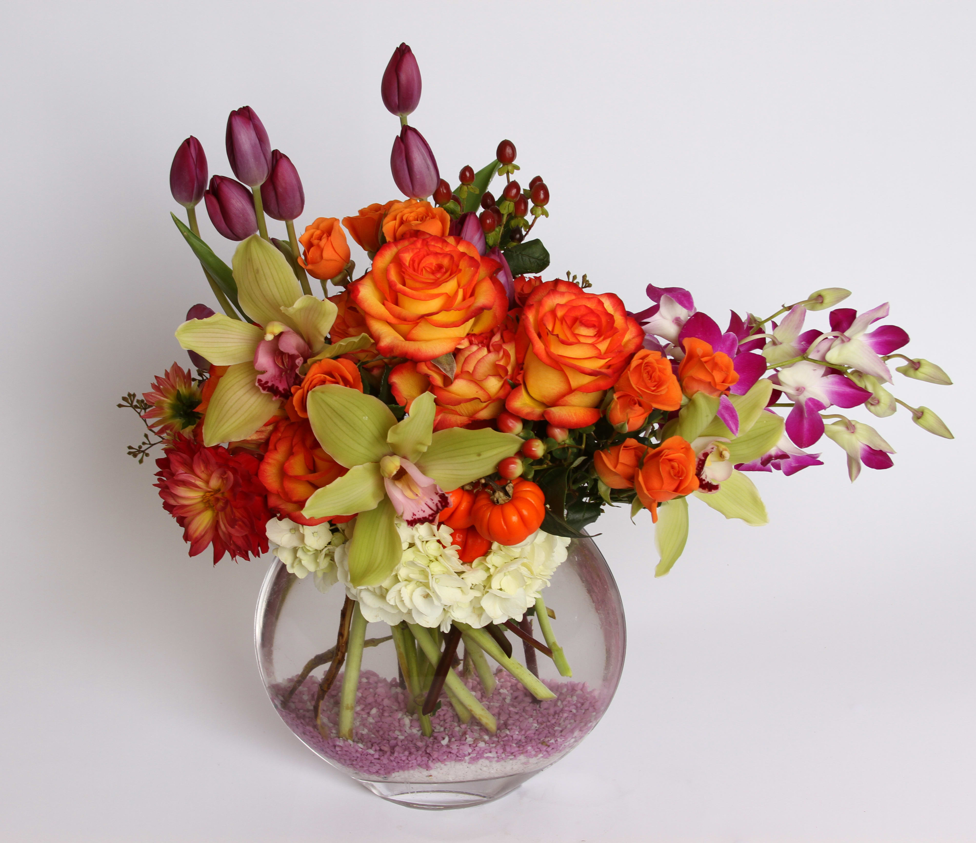 Carnival Carnival - A lively and vibrant bouquet of Carnival roses, purple tulips, dendrobium orchids, cymbidium orchids, dahlias, orange spray roses, hydrangea, hybericum berries and ornamental pumpkins contained in a bubble bowl vase with decorative pebbles
