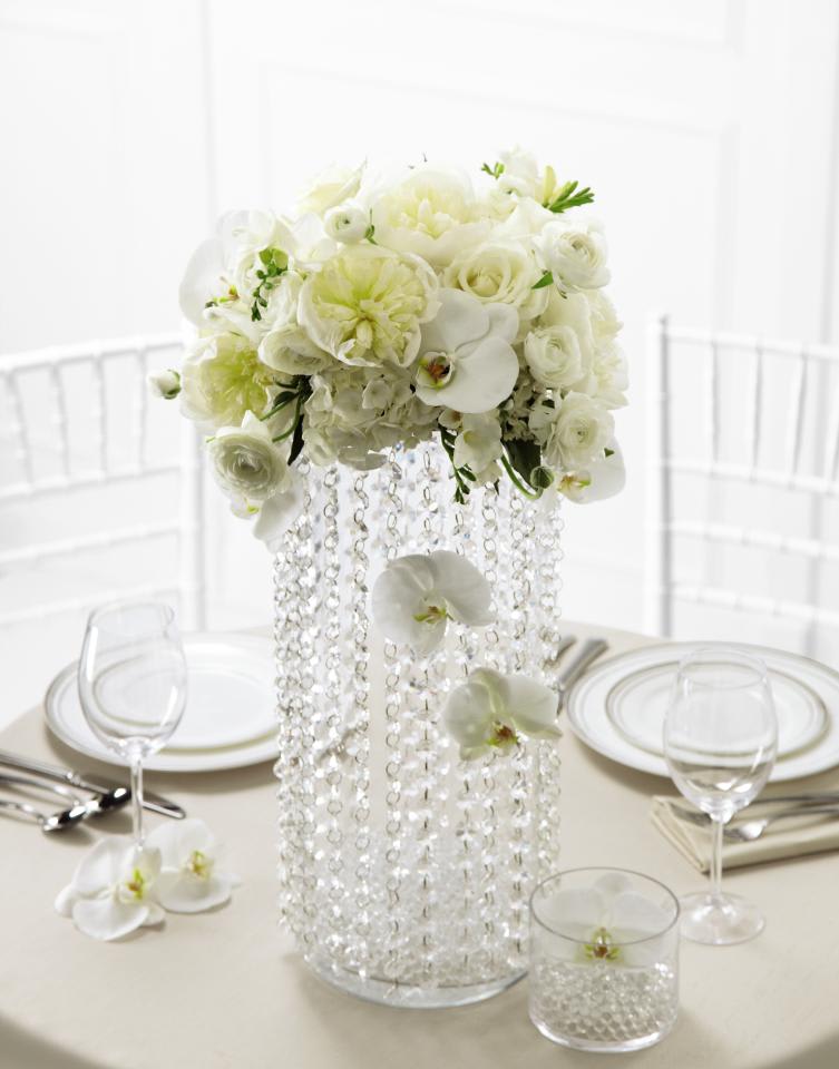 Reception Centerpiece with rental vase - This stunning centerpiece is an eye-catching display of  innovative design and bridal beauty. White ranunculus, peonies, freesia,roses, bouvardia, Phalaenopsis Orchids, and hydrangea are artfully arranged in a cylinder glass vase draped with clear decorative beads to achieve a centerpiece that has a dream-like appeal. Approx. 29H x 14W. 