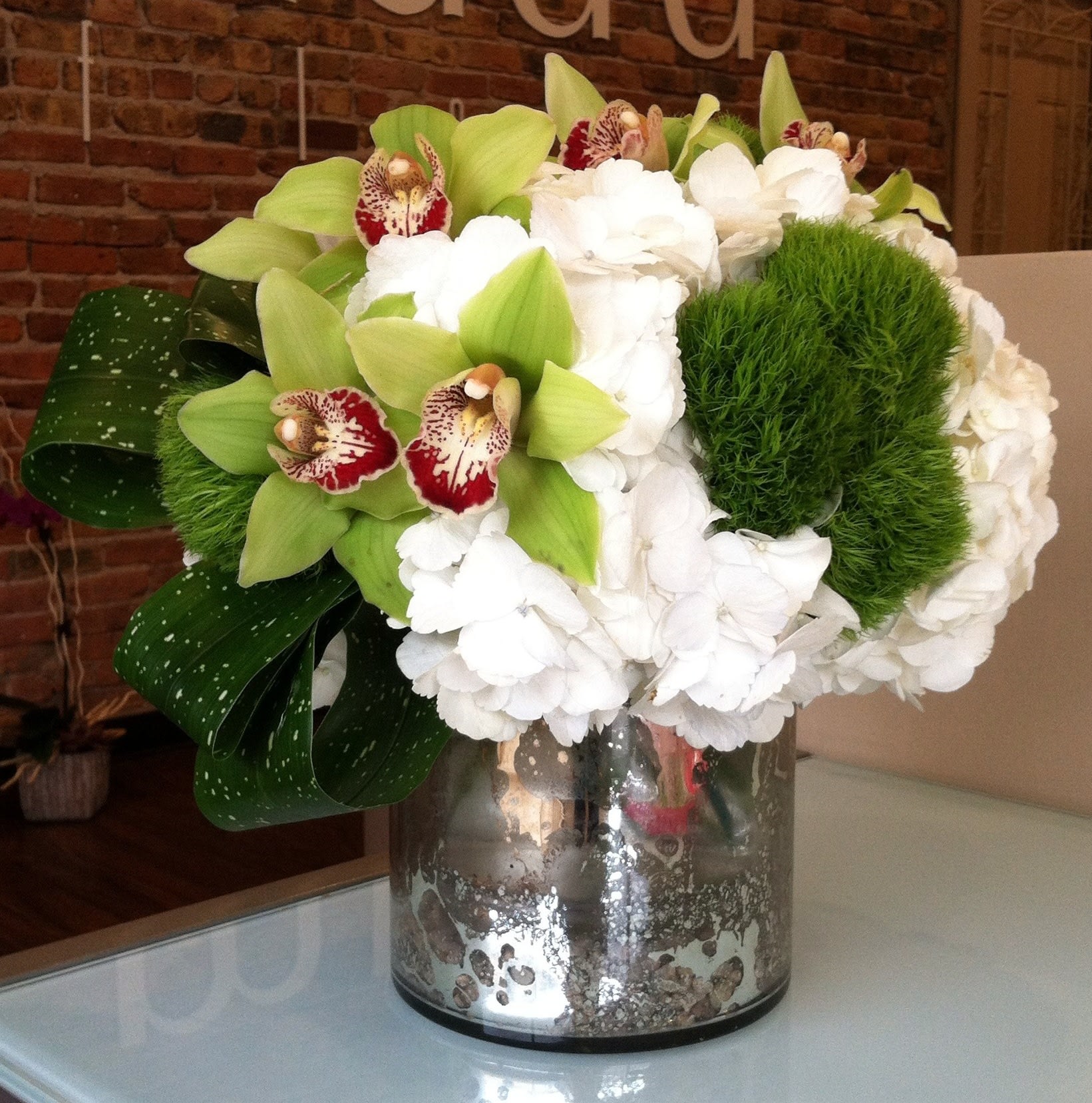 Elegant White and Green &quot;Best of Market&quot; - A designers choice arrangement of white and green flowers that will vary based on market availability in a silver mercury cylinder vase. Arrangement will not be as pictured but in the same style. Pictured is a lush bed of white hydrangea with green cymbidium orchids and dianthus accented by rolled foliage all sitting in a 6x6&quot; silver mercury cylinder.