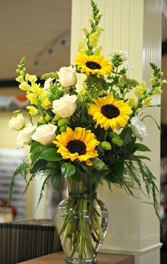 Sun'flower and White Traditions - A traditional bright and bold mix of staple blooms. This artfully created tall piece will have an immediate wow effect in any environment. Sunflowers and Creme'd la creme roses mixed with field greens (minus arugula) will be a choice of good taste.     To prolong the life of this arrangement, please change the water of the arrangement every other day. Call us with questions.  