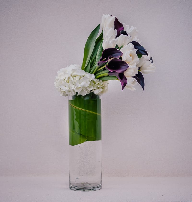 Coutoure - This large and dramatic design features extra long black calla lilies and white tulips set in a tall glass cylinder wrapped with a banana leaf.