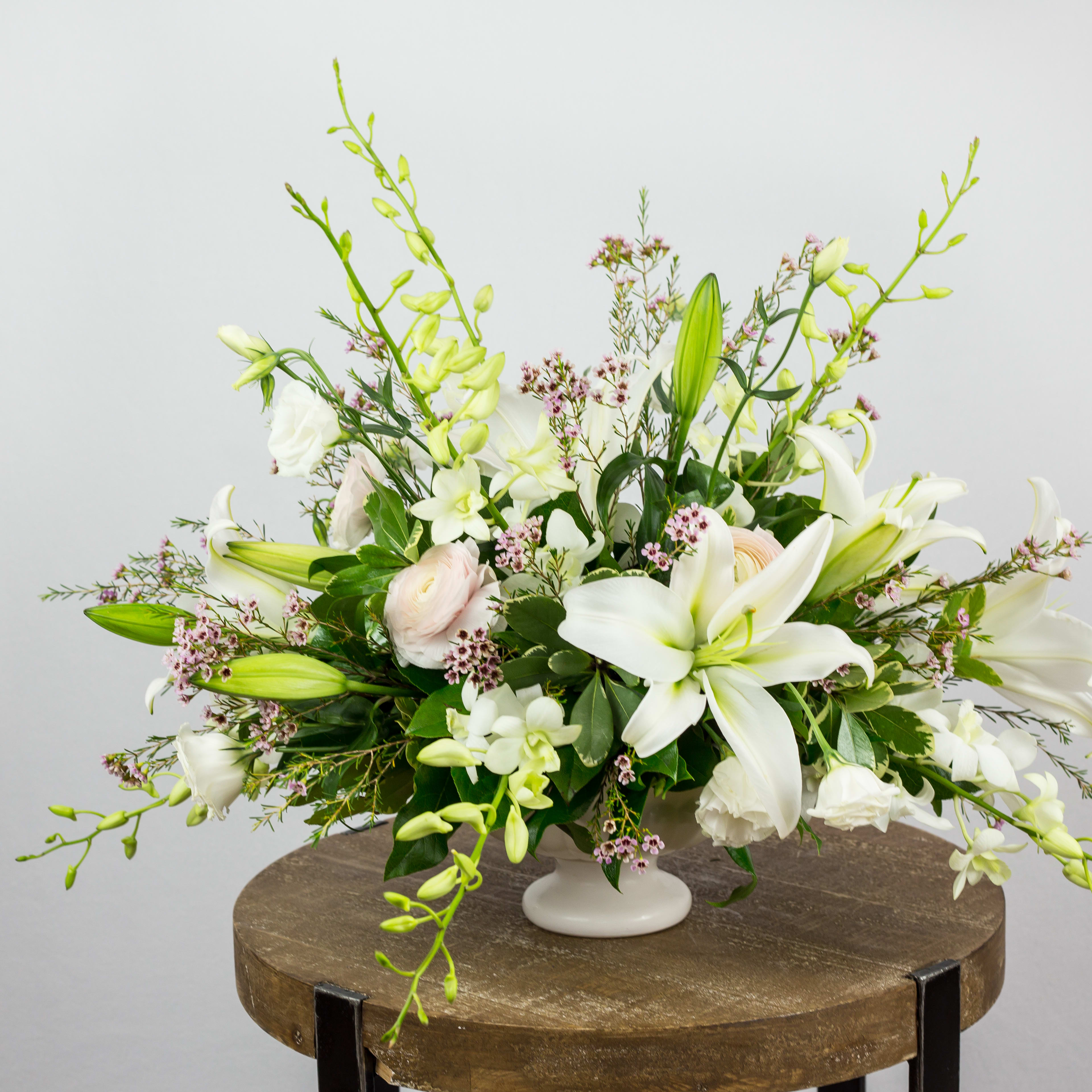 Brightness and Hope - One sided Arrangement in white, footed bowl, Oriental Lilies, Dendrobium Orchids, Lisianthus, Ranunculus, Pittosporum greens. Flowers and containers availability is depending on season and product market.