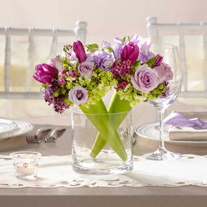 The FTD® Sublime™ Centerpiece -  The FTD® Sublime™ Centerpiece is a presentation of modern grace and elegance. Two clear glass cylinder vases sit side-by-side holding two bouquets in each, set in a diagonal direction for a fresh and sweet look. Each bouquet consists of lavender sweet pea, purple tulips, lavender spray roses, green mini hydrangea, purple lilac, purple waxflower, green hypericum berries and lush greens set in a bright green bouquet cone to give your wedding decor a look of refined sophistication. 