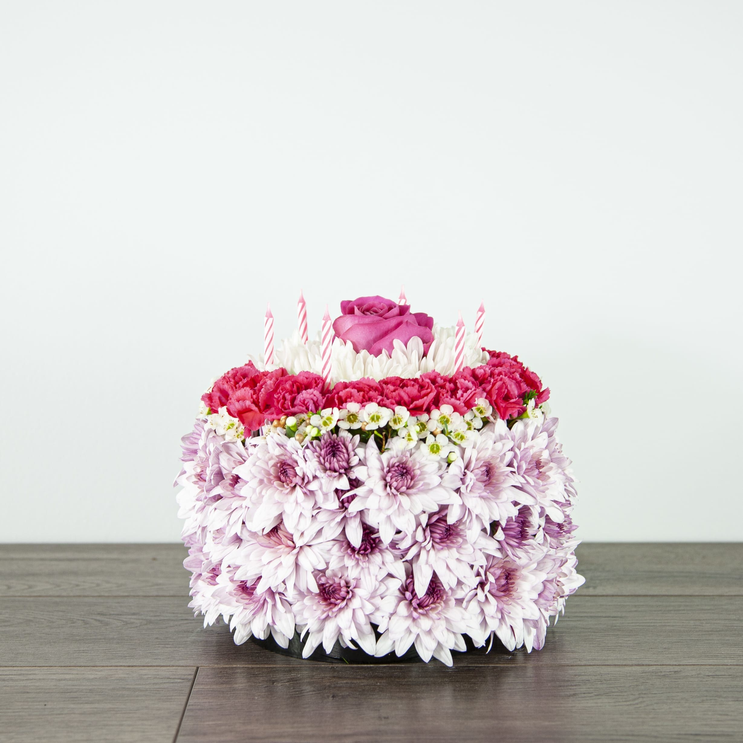 Flower Cake (EXP 60-A) - This celebratory cake is not meant to be eaten, but rather enjoyed by sight. Completed with 6 candles, this flower cake is ready for any birthday celebration.   Blooms in this arrangement: - 12 lavender cushions - 3 white cushions - 1 hot pink rose - 5 hot pink mini carnation stems - 2 white wax flower stems