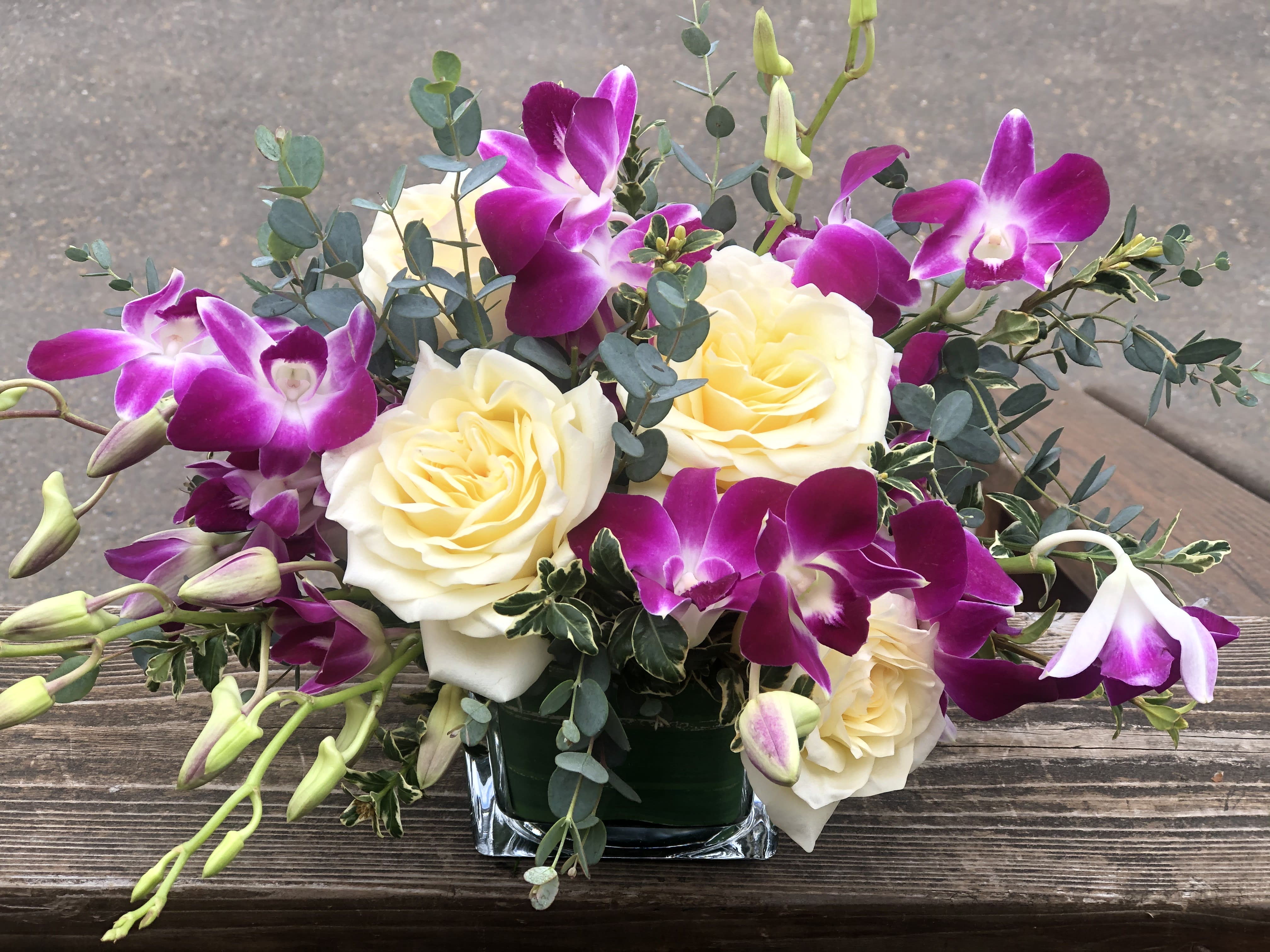Bombay Elegance  - A stylized arrangement  with Bombay orchids, white roses, and mix textured greens in a short cube centerpiece. Our orchid come from Thailand and Hawaii please give us a call if you would like to see about other colors that we may have in. 