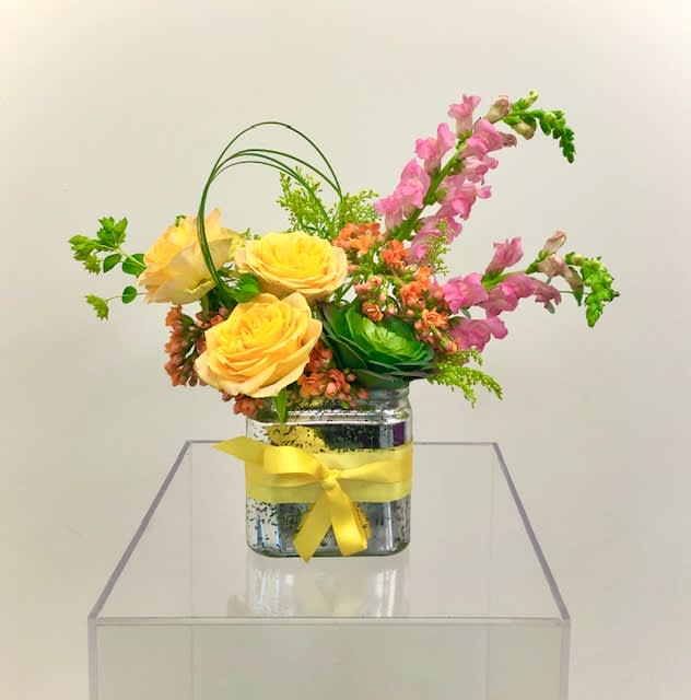 Sassy Suzie - A beautiful design in yellow, pink, and orange tones.  Design consists of roses, snapdragon, succulent, solidago, and various greens to give this arrangement a hint of flair and sassiness.  The silver container can be saved for future use.   *Please view policy section regarding substitutions.