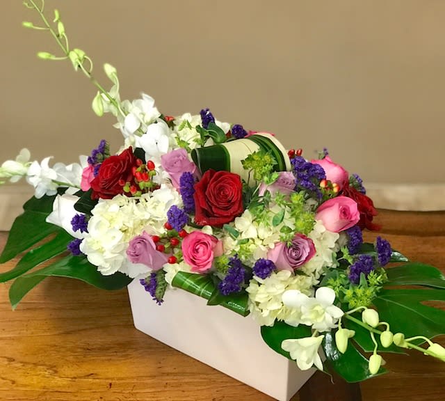 Floral Melody Centerpiece - Beautiful floral arrangement with white and green hydrangeas, red roses, pink roses, lavender roses, white dendromium orchids and ti leaves arrived in a rectangle white ceramic container. 