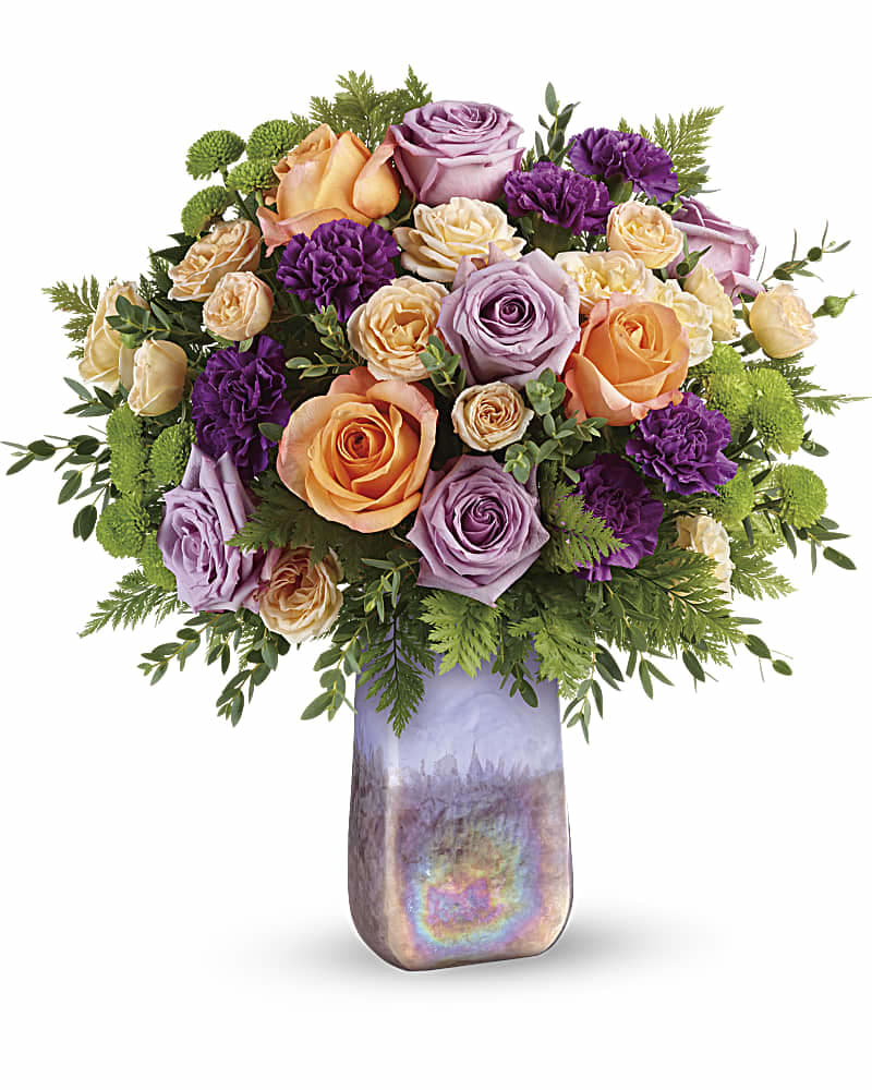 Teleflora's Amethyst Sunrise Bouquet - Two gifts in one! This glorious bouquet of lavender and peach roses is arranged in a hand-blown art glass vase, a shimmering, iridescent gift they'll treasure forever. This glorious bouquet includes lavender roses, peach roses, peach spray roses, purple carnations, green button spray chrysanthemums, leatherleaf fern, and parvifolia eucalyptus. Delivered in an Amethyst Gemstone Art Glass vase.