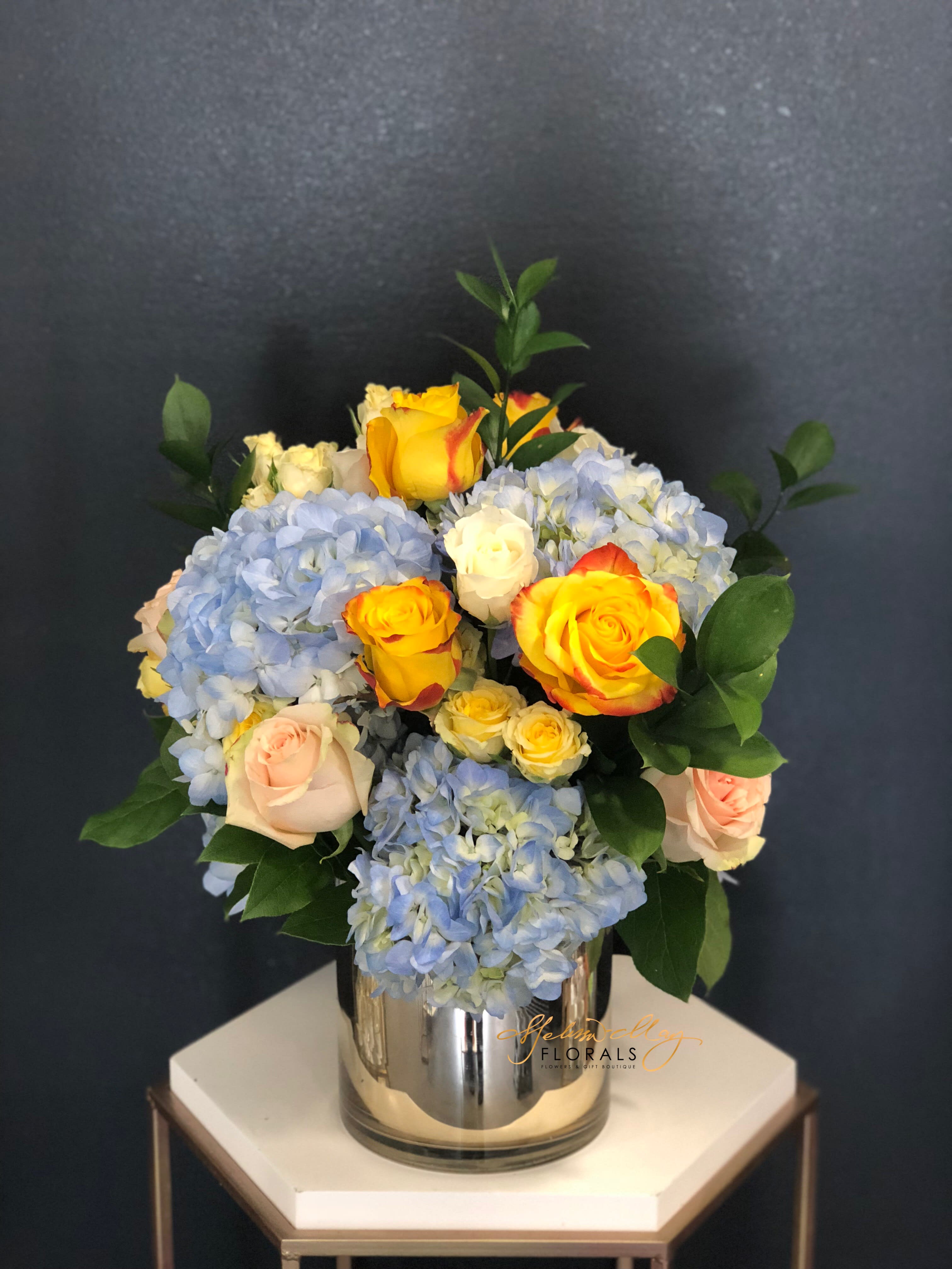Blue Magic - Lets bring home this arrangement to celebrate the birth of your new baby or perhaps the joy of a new beginning. As the blue hydrangeas brings out the entire arrangement with those orange roses popping right at you. Lets not forget about those blush roses to finish off the arrangement.  