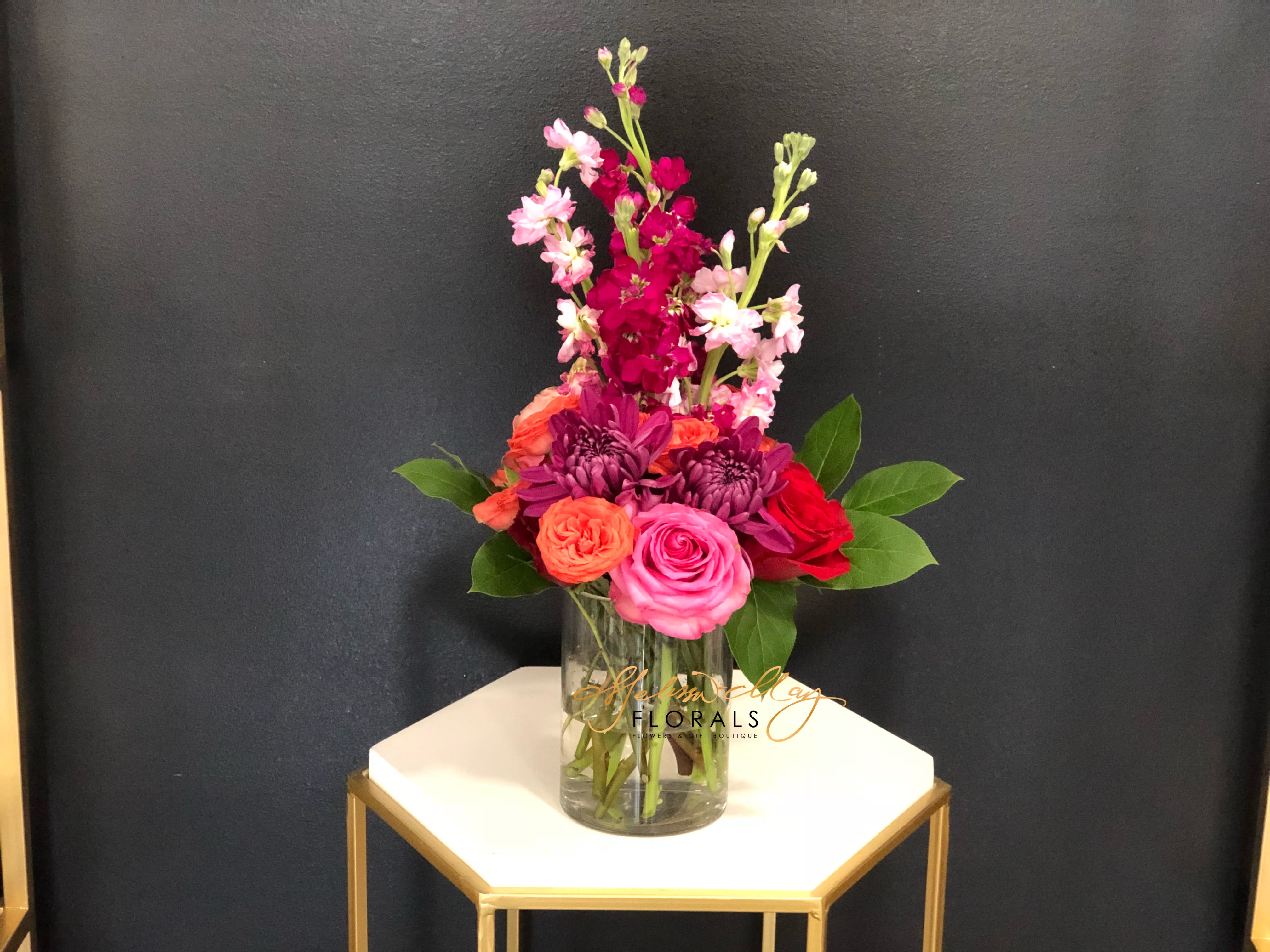 Bright Arrangement - Here you see pink and purple stock stems, orange spray roses with lush greenery. An all around bright arrangement that anyone would fall in love with. Its nothing like having a bright arrangement that will light up an entire room. 
