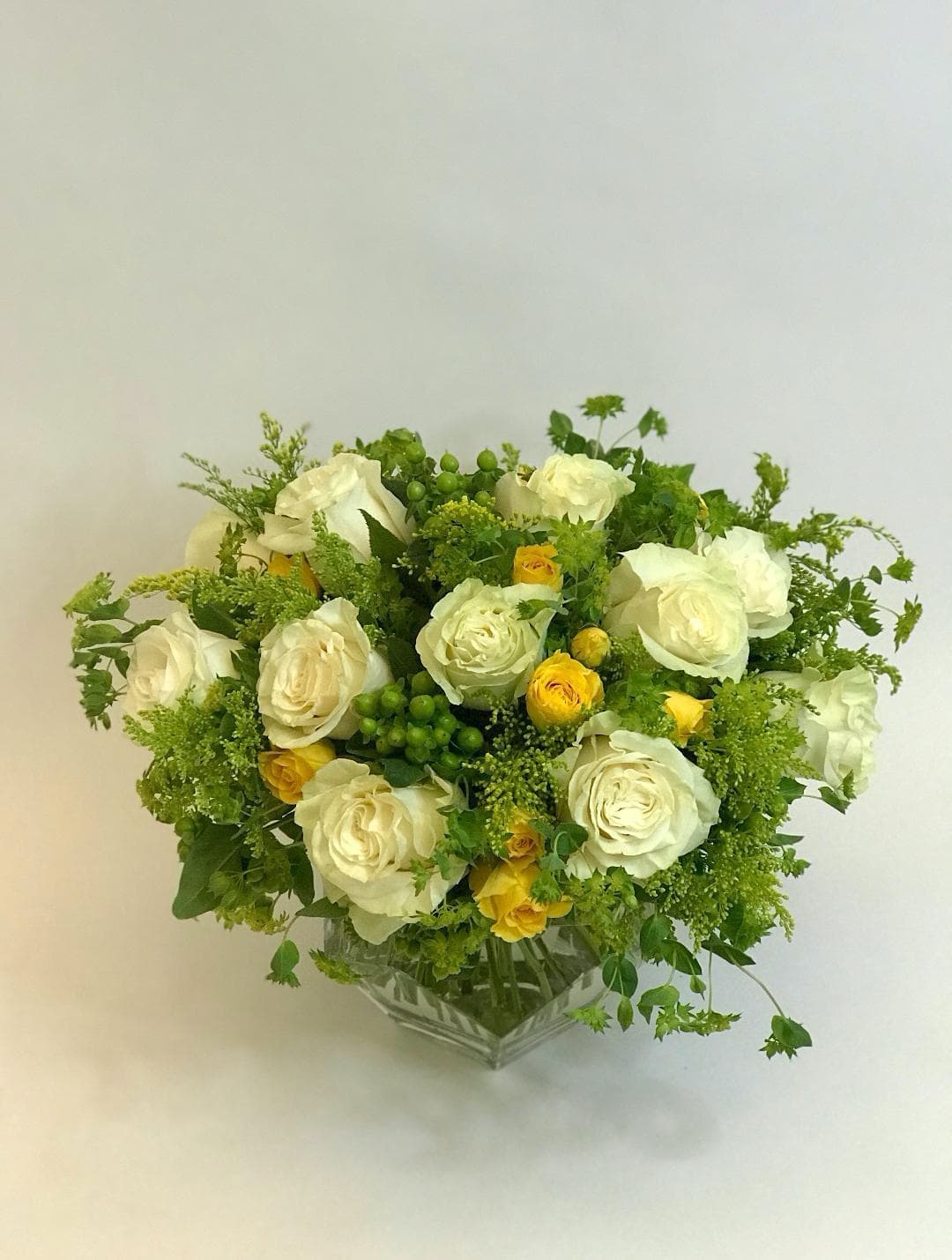 golden solstice - Beautiful fresh blooms of Mondial white roses, green coffee bean, yellow spray garden roses, blueplurum, and solidago in geometric glass vase