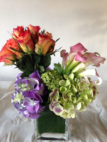 Bright and Beautiful - Vibrant orange Roses, pink Calla Lilies and lavender Hydrangeas bunched and displayed in various angles, and placed in a tall clear glass container.  Comparable substitutes will be used for seasonal flowers.