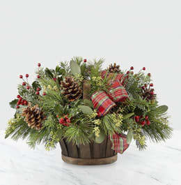 Holiday Homecomings Basket - Whether you're welcoming your family home or sharing a gift filled with natural cheer, our Holiday Homecomings Basket is the perfect arrangement to do so. A lush collection of assorted Christmas greens, seeded eucalyptus and holly come together a rustic wooden basket. Accented by a red and green ribbon, pinecone pics and metallic berry pics, this arrangement adds a touch of charm to wherever its placed. 