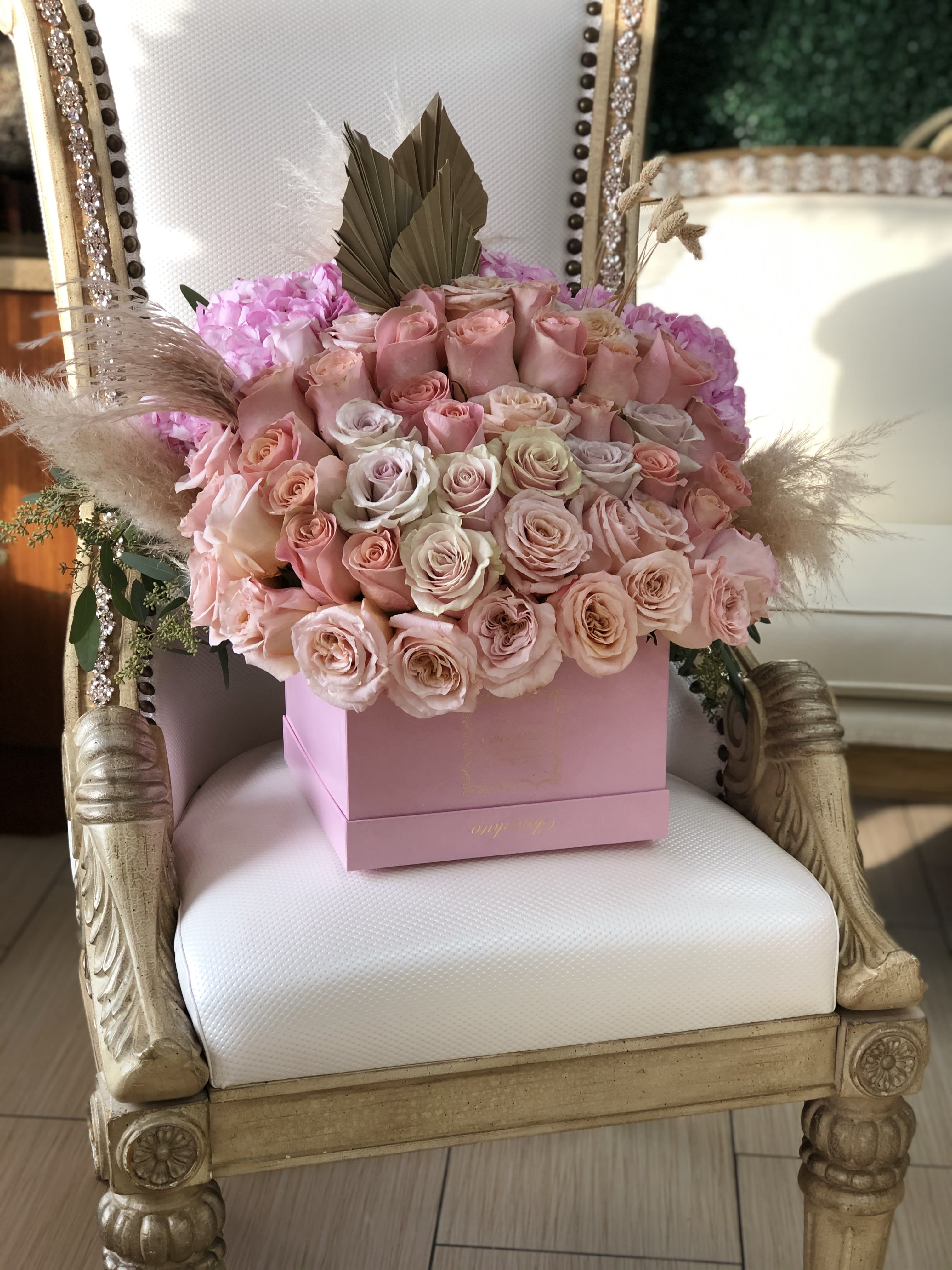 Penelope - Pink Roses with Pampas Penelope - Pink Roses with Pampas - This Chocolato arrangement is made with Pink Roses with Pampas. The arrangement comes in a pink sqaure box. Please see care instructions for further assistance on taking care of your Chocolato Boxed Flowers. All prices are subject to change due to season and availability of fresh flowers. The arrangement seen is what will be made unless any substitution may need to be made based on season and availability of flowers. Box shape and color might differ based on availability.