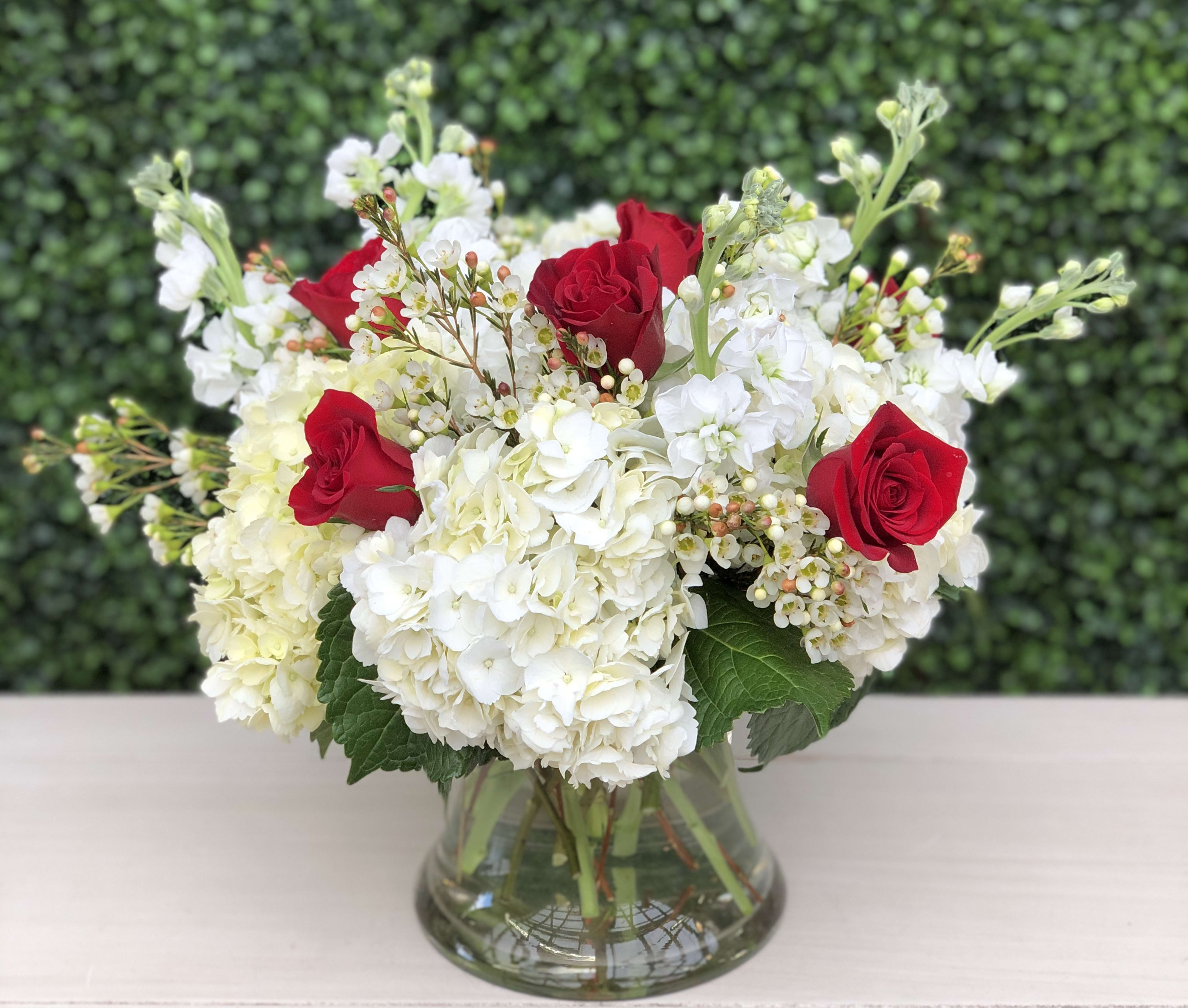 Full Of Romance - A compact arrangement of white hydrangeas, red roses, white stock and white wax flower in a clear glass gathering vase. 