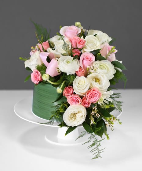 Blooming Pastel Cake - This flower cake will cause oohs and ahhhs. A lovely combination of garden roses, ranunculus, pink spruce, waxflower, and assorted greens are served on a cake plate to celebrate that special occasion. (Due to availability some flowers will be replaced with equal or higher value colors will be respected).