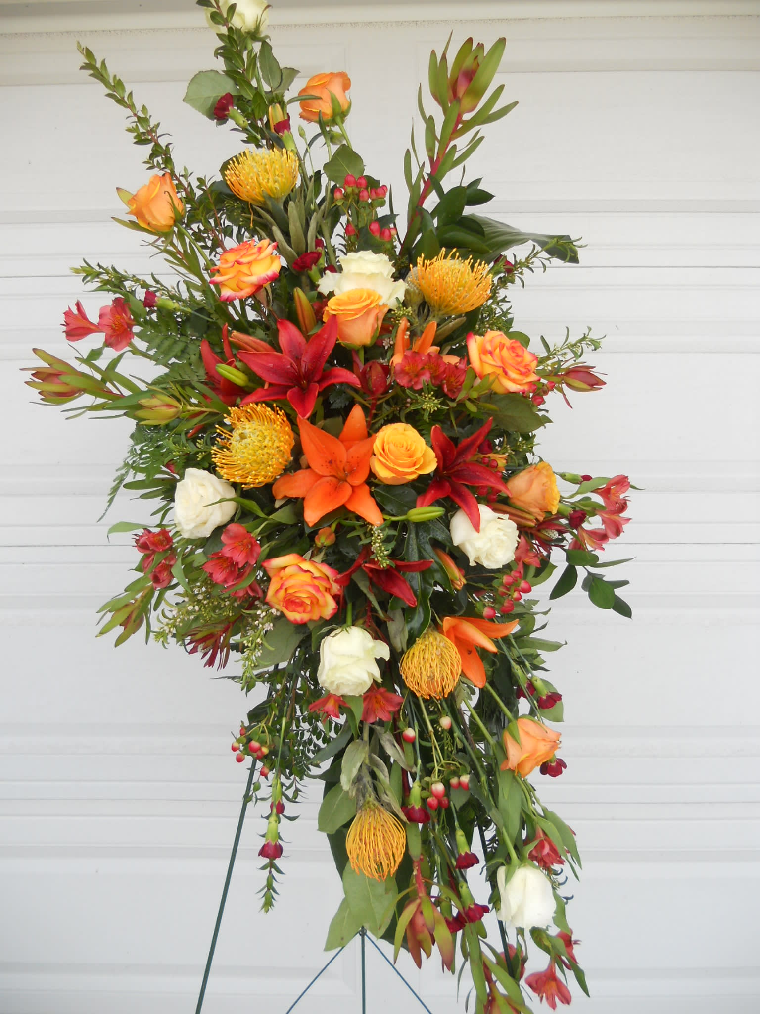 Masculine Orange, Yellow, Red and White Easel Spray Masculine Orange, Yellow, Red and White Easel Spray - This Tribute for a man features Pin Cushion protea, lilies and roses. Also adding to the design are alstromeria, berries a yellow filler flowers.