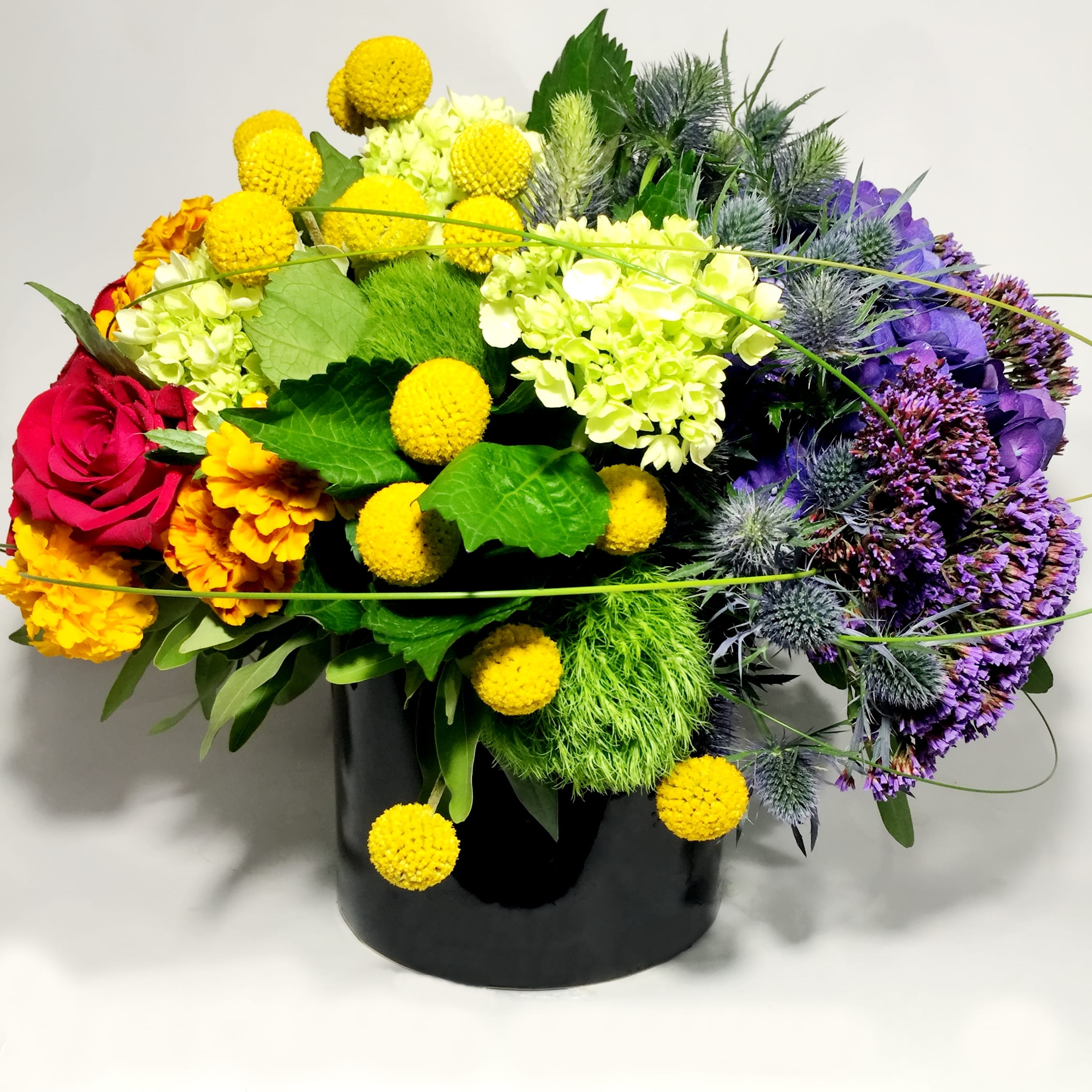 Loud And Proud Vase Arrangement - If you need to send someone some rainbow love or show your unending support this bountiful bouquet is a great way to do it. Covering all the colors of the rainbow the floral collection may include orange marigolds (subject to seasonal availability) Red Roses, Green Hydrangea, Green Trick Dianthus, Yellow crespedia, Blue Thistle and Purple Statice. Everything is accented by sweeping blades of grass and perfectly mounded together in a sharp cylinder vase, it's certainly a show stopper.  Measuring Approximately 16&quot; by 14&quot; *Due to availability, on occasion it may be necessary to substitute a certain flower or container for your order.