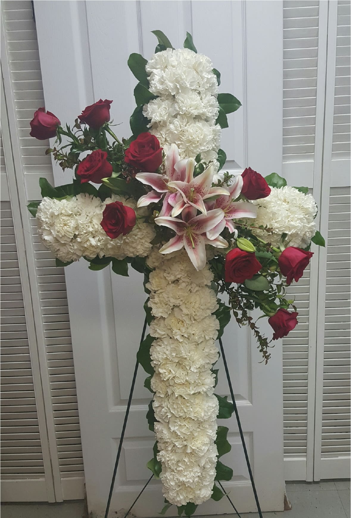 White Cross Standing Spray - Commemorate fidelity with this standing spray cross of white blossoms adorned with fragrant lilies and fresh red roses. It’s a display that remembers faith, hope and charity. 