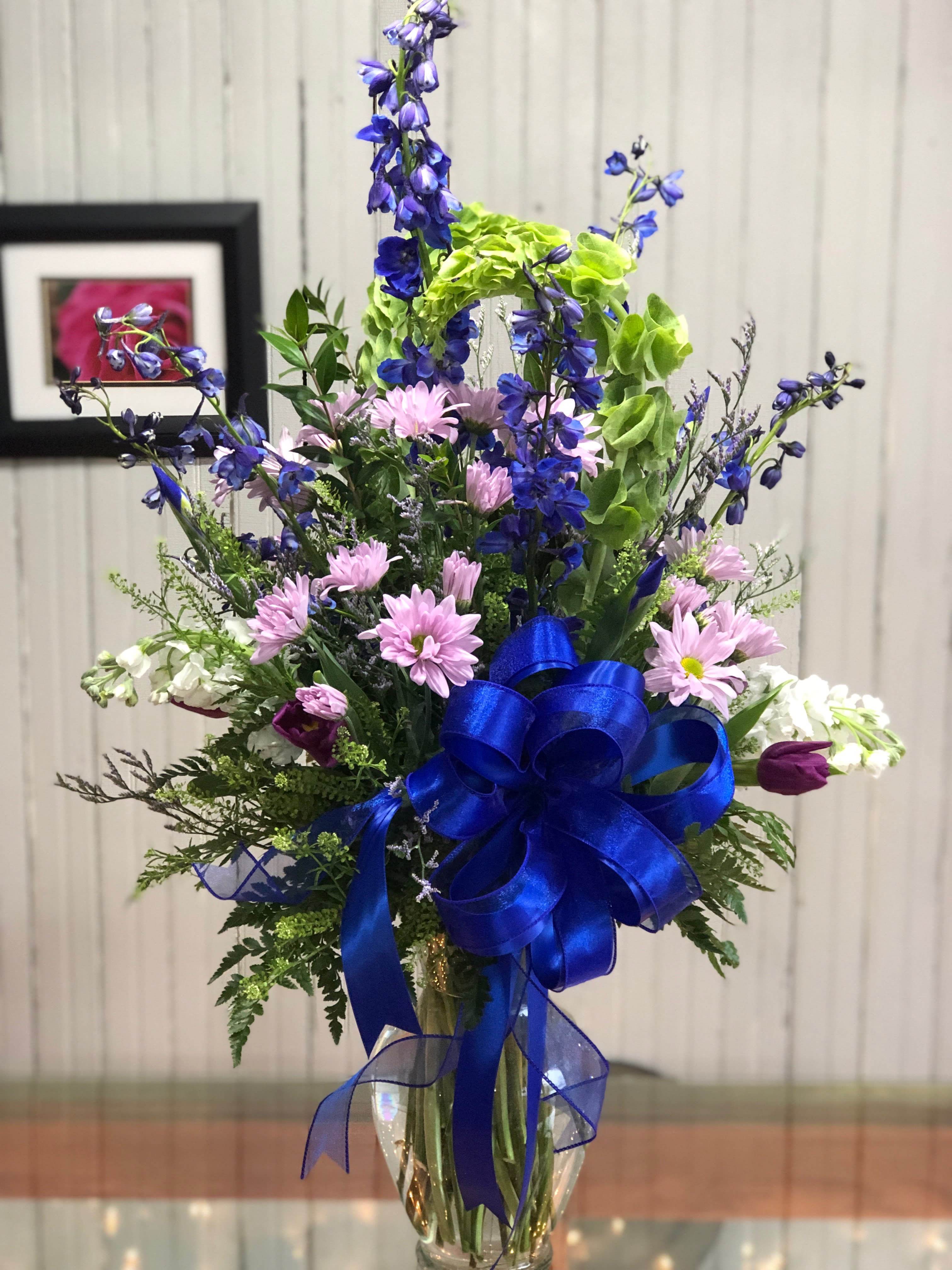 Remember Me - This is a grand arrangement of blue delphinium, bells of Ireland, and purple daisies and accents, in a clear glass vase.   ****We will try our best to match the flowers in the picture, but there may be times when we substitute flowers.  We will always try to make it have the same look and feel as the original.
