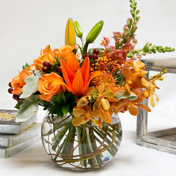 Orange Sunset AD10 - This incredible arrangement mirrors the colors of the sunset. Roses, coffee bean, protea, tropical flowers and other seasonal flowers and greens are artfully arranged a glass Bubble-Bowl with a curly willow wrap.