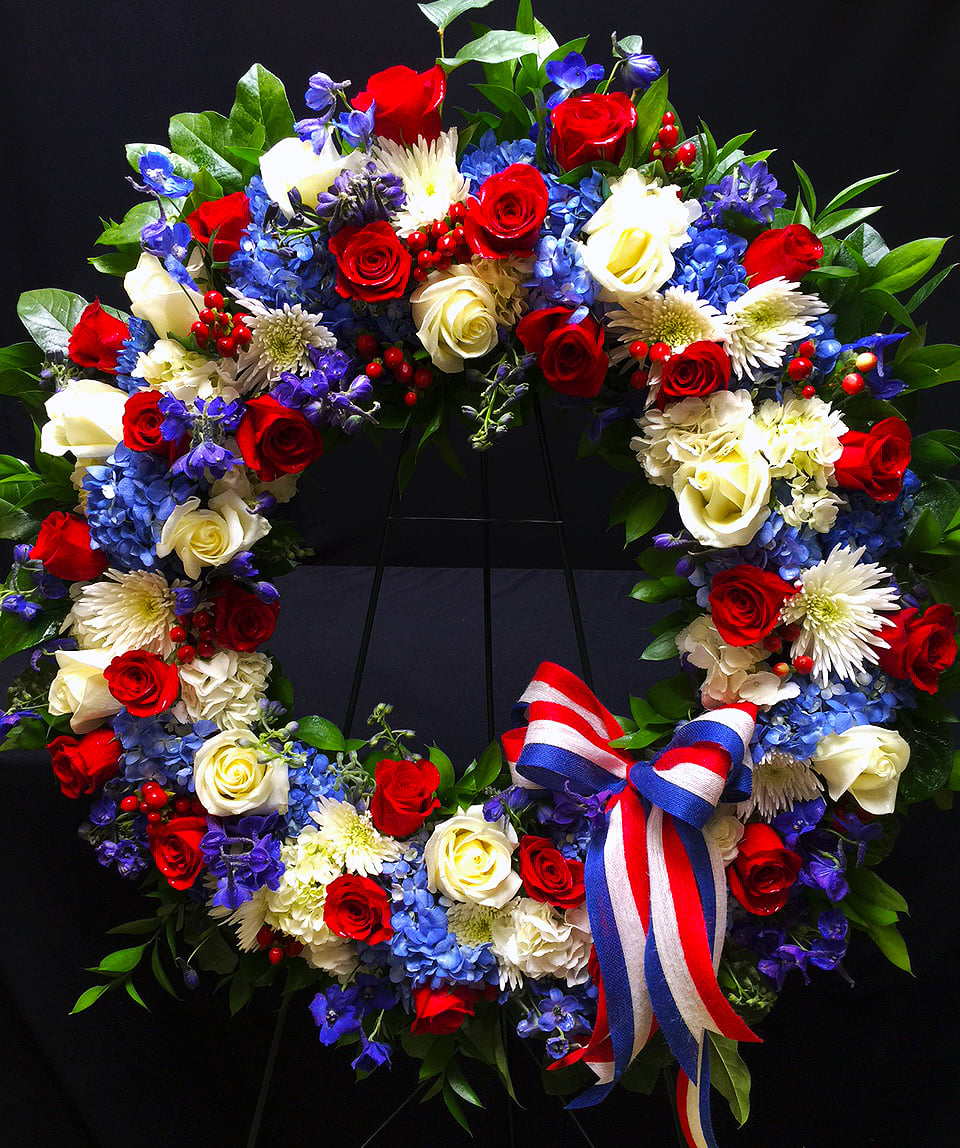 American Tribute AF10 - This Patriotic Wreath is paved with seasonal flowers, hydrangea and roses in Red, White and Blue. It is finished off with red, white &amp; blue ribbon and presented on a 5' tall metal easel.