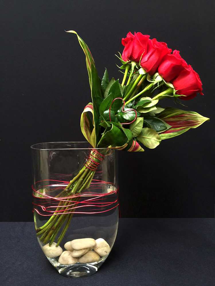 Heart Song V15-6 - Why always send the same old traditional red roses and baby's breath? After all it isn't 1952 any longer! Roses are still a sign of romance and love so Adelaide's created a new look for a dozen long-stemmed roses circa 2014 and beyond. Set in a tall envelope vase, filled with white rocks, the roses and vase are bound with red wire and accented with a red wire heart. Perfect for the Valentine in your life to say &quot;Yep, I'm cool&quot;.