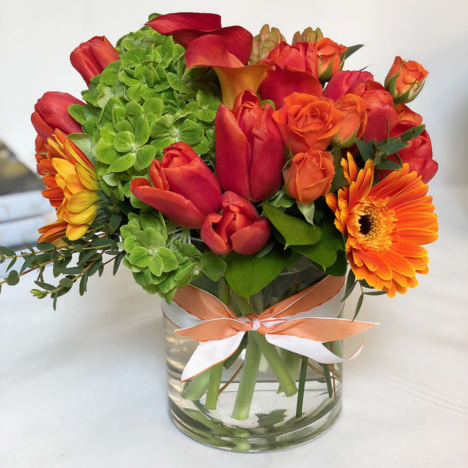 Fun Loving Orange MD18-3 - The perfect arrangement for that fun loving person in your life. Tulips, Ranunculus, miniature calla lilies and other season flowers are artfully arranged in a clear cylinder vase with a fun ribbon tie. 