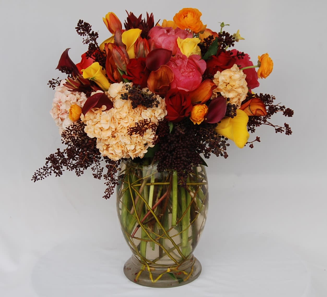 Dressed For Autumn  - A vase filled with hydrangeas, coral peonies, gold, orange, burgundy calla lilies, orange tulips, red roses, and orange Ranunculus, accented with seeded eucalyptus and curly willow.