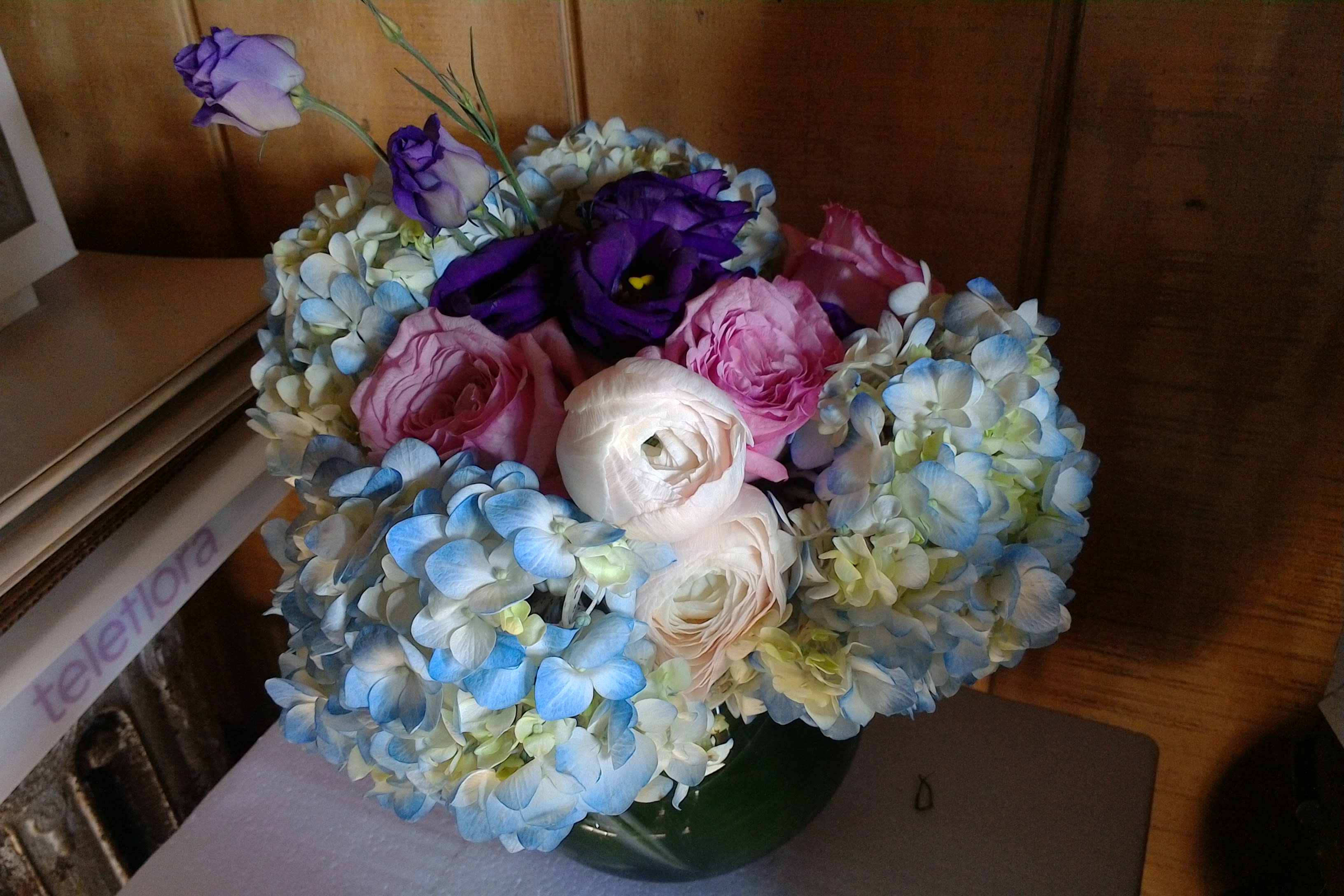 Sweet Love - A beautiful floral arrangement of blue hydrangea, blush pink ranunculus, pink spray roses and purple lisianthus in a round clear bowl.  This arrangement can be made in different colors upon availability.
