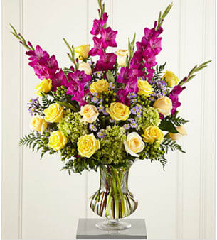 The FTD® Loveliness™ Arrangement - Big, colorful, sophisticated. This generously-scaled arrangement makes a fresh and loving tribute to a life lived in beauty and grace. A superb choice for a wake or a funeral service, this colorful bouquet is handcrafted by a local FTD artisan florist of yellow and cream roses, fuchsia gladiolus, lavender monte casino, green hypericum berries and hydrangea with accents of greenery in a premium glass vase. You can count on its making a comforting impression that will be long-remembered.  Your purchase includes a complimentary personalized gift message.