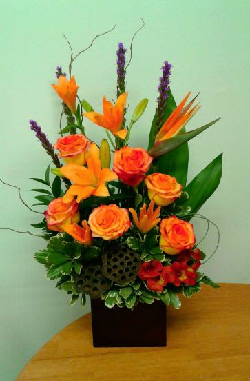 Colorado Sunset by Floral Expressions - A dramatic floral design inspired by the spectacular colors of a Colorado sunset.  Featured in this arrangement are Orange Asiatic Lilies, Orange Roses, Tropical Birds of Paradise (when seasonally available), Orange Alstroemeria and Purple Liatris.  Arranged tall in a large wooden cube, accented with curly willow, lotus pods and unique foliage.  