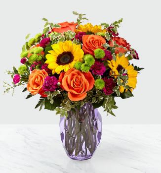 Summer Craze Bouquet  - Our floral professionals beautifully combine a blend of orange roses, sunflowers, violet mini carnations, green button poms, and lush greens to create this stunning arrangement.  Presented in a modern geometric purple glass vase, this beautiful design is available for same day delivery to Washington Park, Cherry Creek, Belmar and most Denver locations by Floral Expressions, Denver's Best Florist!  