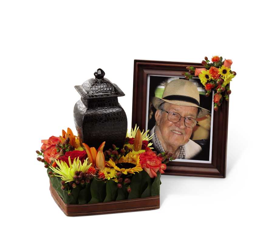 The FTD Glorious Tribute Arrangement - The FTD Glorious Tribute Arrangement is a beautiful way to honor the life of the deceased. Orange bi-color roses, red roses, orange safflower, viking bi-colored chrysanthemums, yellow spider chrysanthemums, sunflowers, orange asiatic lilies and lush greens are arranged on a sculpting sheet accented with a copper decorative wire and chocolate brown ribbon in which the urn is placed towards the center, creating an incredible display of love and light at their final tribute.