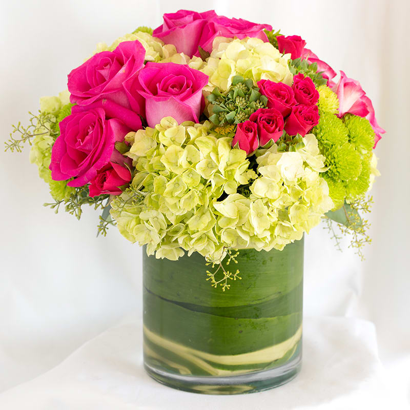 Simply Gorgeous - Modern and chic, clusters of roses, spray roses, hydrangreas put together in a compact, all-around arrangement. Cylinder vase has Ti-leaf wrap to mask the stems and add elegance. 