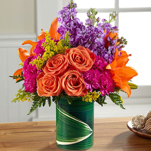 The FTD® Sunset Sweetness™ Bouquet - Take a quick and fun spin around the color wheel with a bouquet of bright, vibrant shades spanning a sunset spectrum from orange through purple. Orange roses (in Better, Best, Exquisite), Asiatic lilies, Gerbera daisies (in Good only), magenta carnations, purple stock and more are arranged in a ceramic &quot;illusion&quot; vase that convincingly replicates the look of a leaf-lined glass pillar. It's an arrangement sure to make some lucky someone's day while burnishing your credentials as a most creative gift giver. BETTER bouquet is approximately 14&quot;H x 12&quot;W. 
