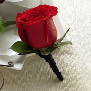 Red Rose Boutonniere Red Rose Boutonniere - The Red Rose Boutonniere creates the classic picture of wedding elegance. A single red rose is chosen for its absolute perfection then accented with lush greens and a black ribbon to offer its beauty and sweet fragrance in honor of the celebration. Contact us for custom orders!