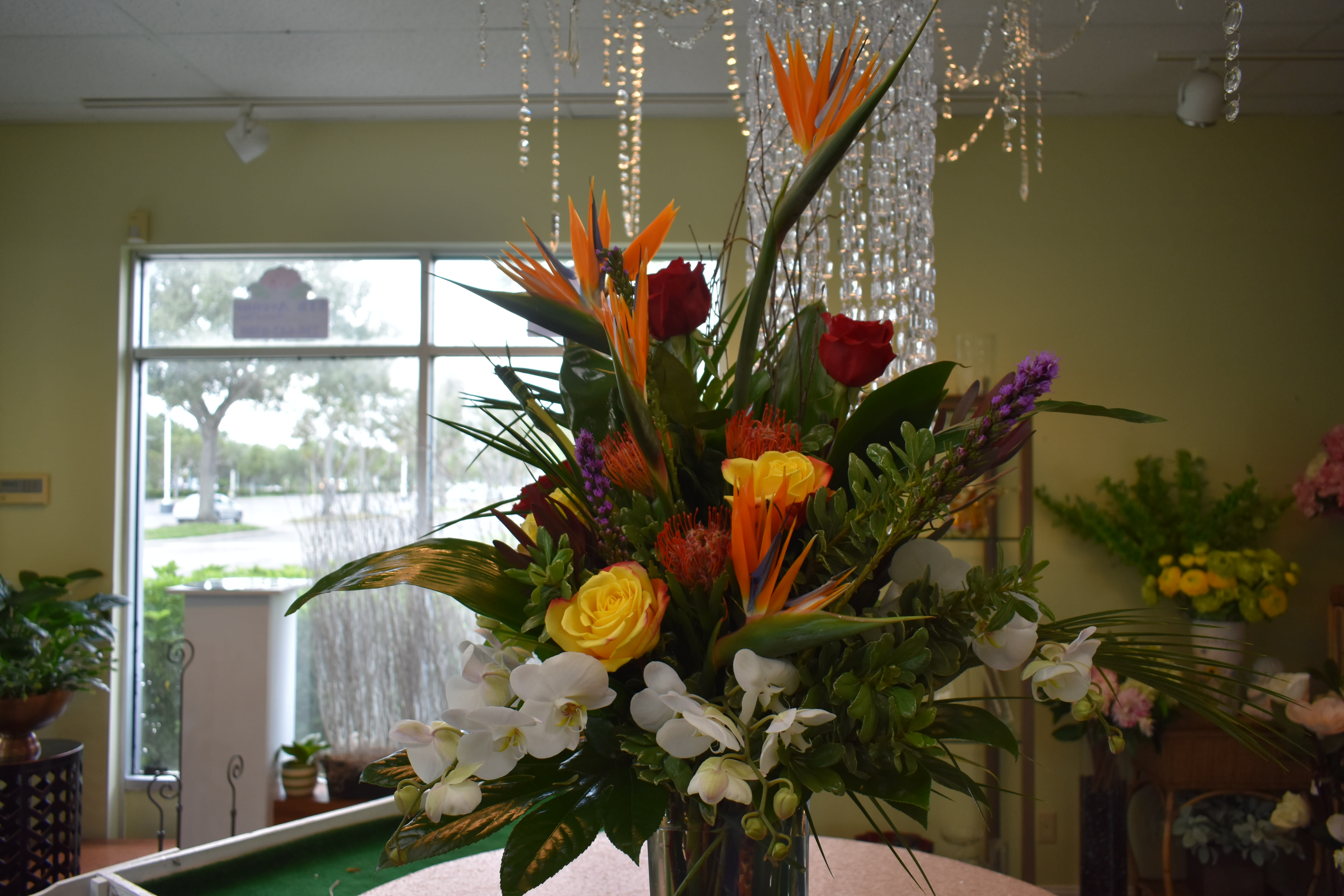Tropical Paradise  - Birds of Paradise, Red Roses, Yellow Roses, White Phalaenopsis, Tropical Greens with Curly Willow Wrapped in green leaf in tall Elegant rectangular vase