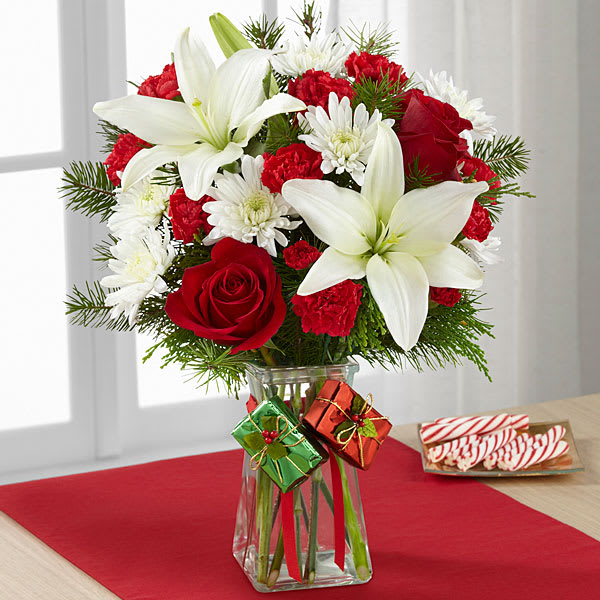 The FTD Joyous Holiday Bouquet - The FTD® Joyous Holiday™ Bouquet is a seasonal sensation your special recipient will love. Rich red roses carnations and mini carnations are brought together with brilliant white Asiatic lilies and chrysanthemums accented with lush holiday greens to create an incredible fresh flower arrangement. Presented in a clear glass gathered square vase embellished with red ribbon at the neck dangling foil-wrapped present accents to give it a festive look this holiday flower bouquet will make an excellent Christmas gift for your favorite friends family or co-workers.