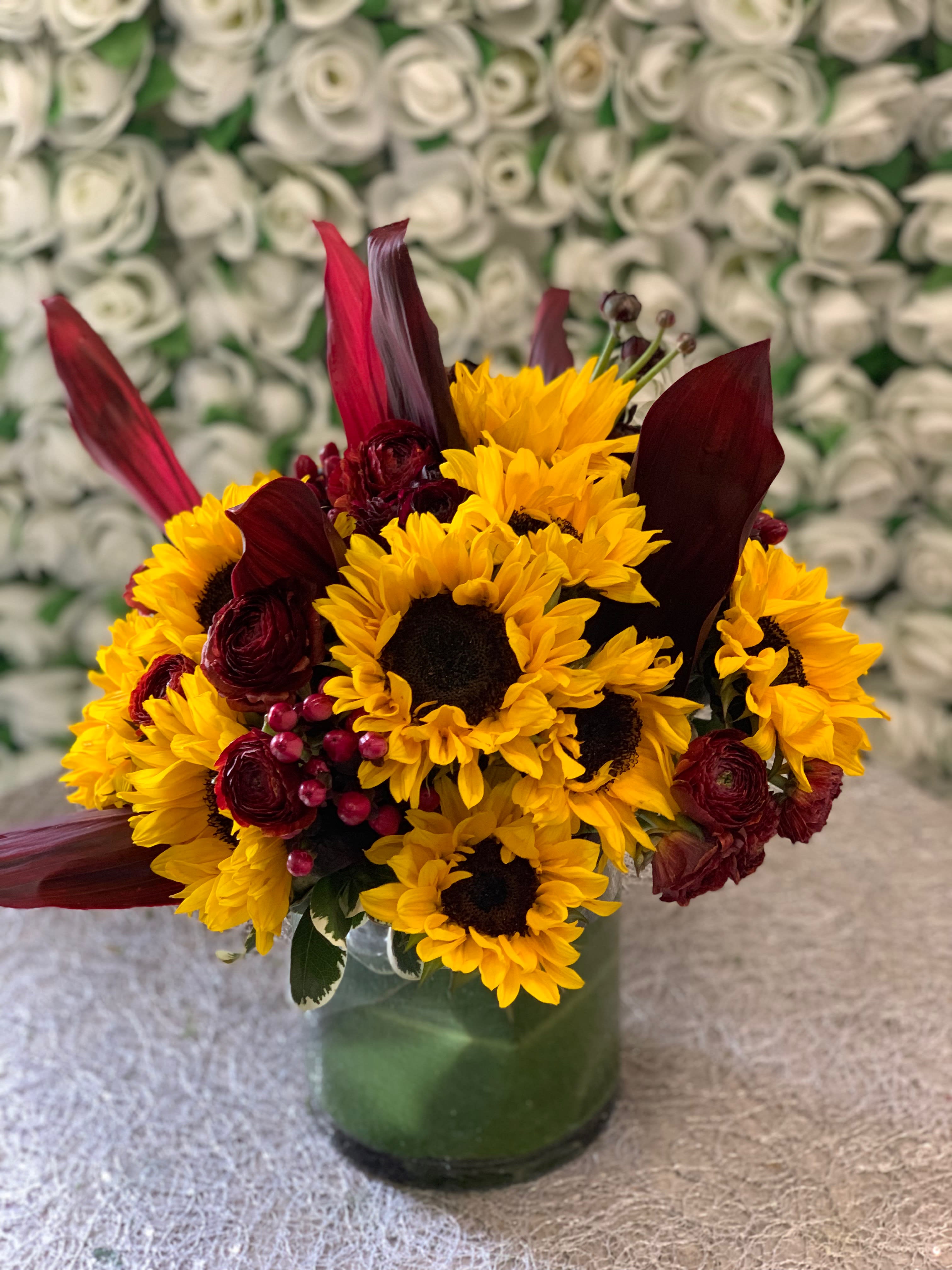 Sunshine Delight - Bunch of sunflowers combined with ranunculus and coffee bean6 gathered in a cylinder vase with red leaves.