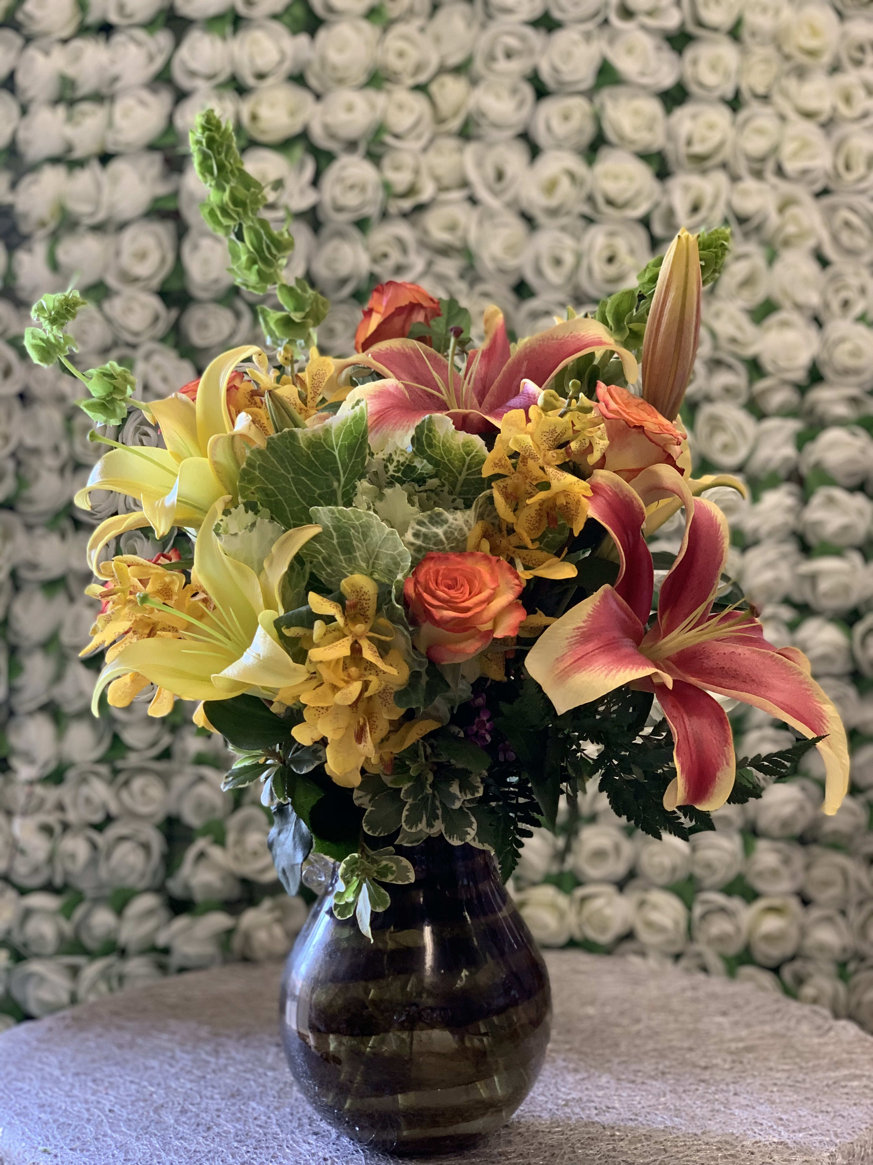 Autumn Aroma - ****PLEASE CALL TO CHECK IF WE HAVE THE EXACT VASE (VASE SHAPE MAY VARY)*** An assortment of elegant lilies and orange roses intertwined with beautiful yellow mokara orchids in a pot gathering vase. 