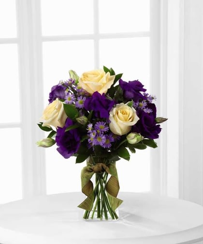 Angelique Bouquet - Create a special moment for someone special.  Cream roses are arranged to perfection against the royal purple hues of double lisianthus and Monte Casino asters and are perfectly accented with lush greens. Presented in a clear gathering square glass vase and tied at the neck with an amber wired taffeta ribbon.  This bouquet is an exquisite way to extend your warmest wishes and affection.